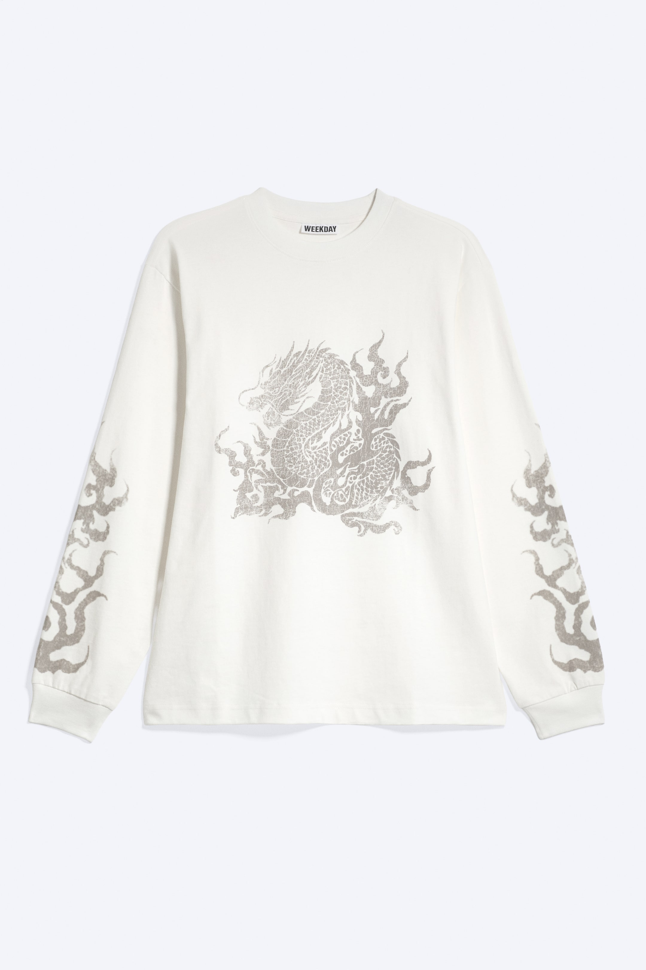 Visa större bild: En krämfärgad långärmad T-shirt visas framifrån med avslappnad passform, ribbstickad rund halsringning, lätt sänkta axlar och ribbstickade ärmslut. Plagget har ett gråtonat grafiskt tryck av en drake omgiven av lågor på bröstet, med matchande flam-mönster längs båda ärmarna.