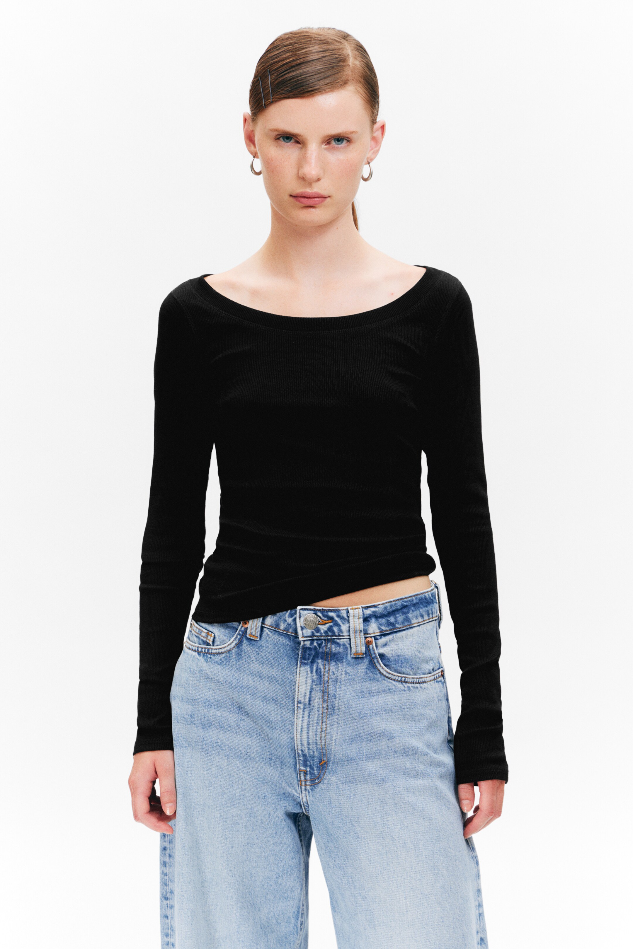 Größeres Bild anzeigen: Ein schwarzes, eng anliegendes Langarm-Top wird getragen, das einen breiten Rundhalsausschnitt und eine gerippte Textur aufweist. Es wird mit hell gewaschenen blauen High-Waist-Jeans kombiniert.