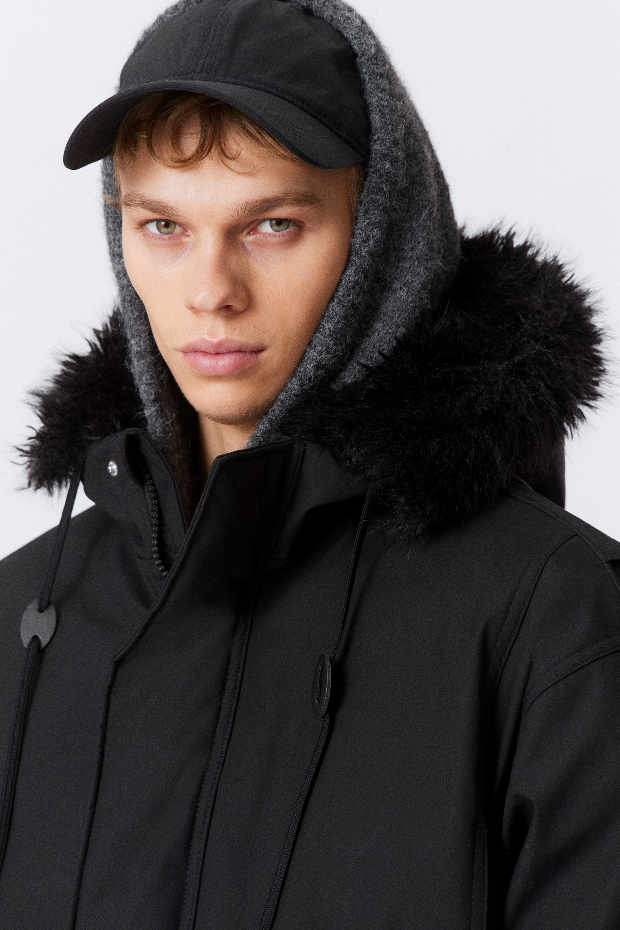 Grotere afbeelding bekijken: Een zwarte parka wordt gepresenteerd, met een hoge kraag met een zichtbare rits en zwarte namaakbont rand rond de kap. Het is gestyled met een getextureerde grijze capuchon die eronder is gelaagd en een zwarte baseballpet.