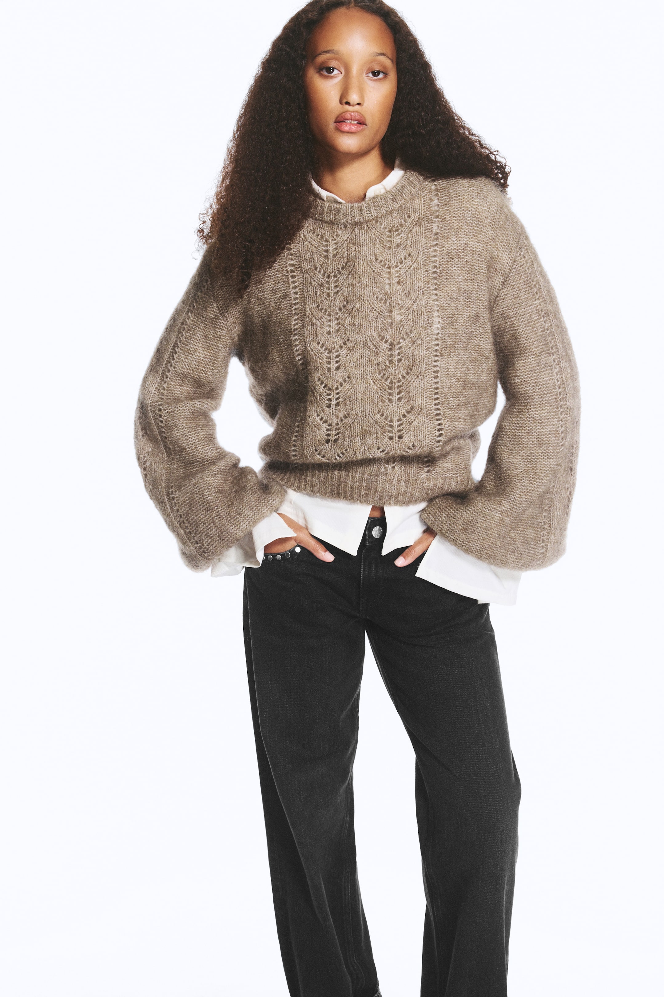 Größeres Bild ansehen: Ein Pullover in Taupe aus Mohair-Mischung mit Rundhalsausschnitt, dekorativen Lochstrick-Einsätzen entlang der Vorderseite und weiten Ballonärmeln wird vom Model getragen. Der Pullover ist über einem weißen Hemd mit Kragen getragen und mit einer schwarzen Jeans kombiniert, während das Model mit den Händen in den vorderen Taschen steht.
