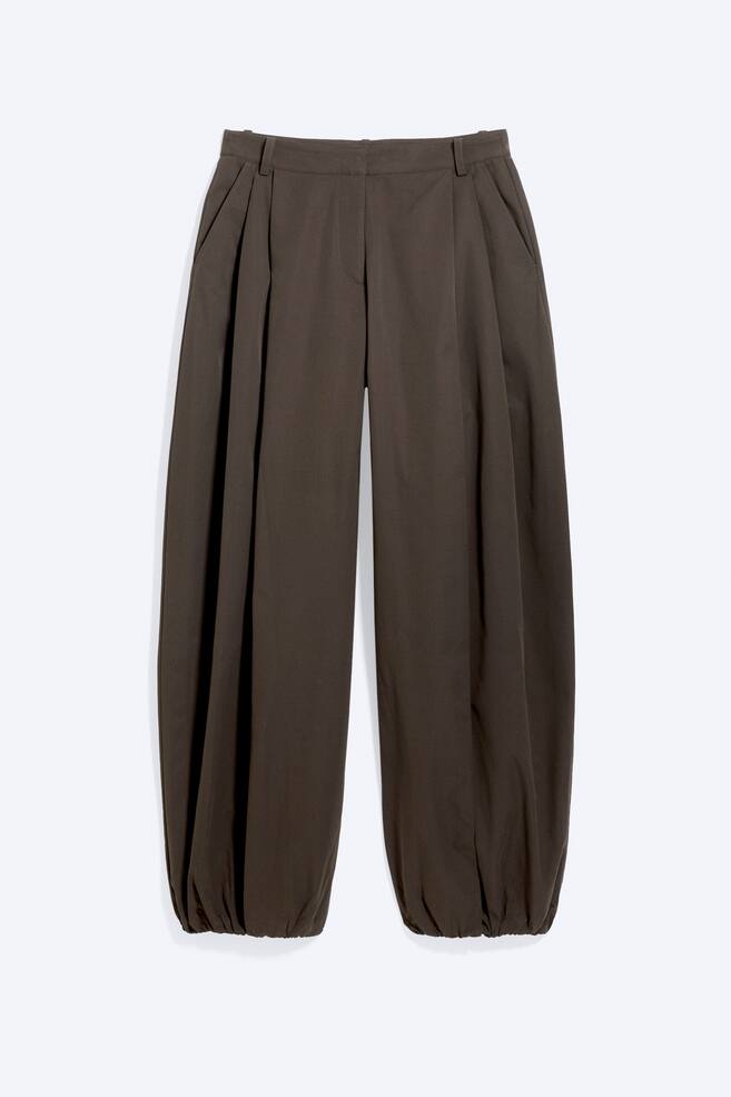 Die dunkelbraunen Hosen präsentieren sich in einer Vorderansicht und zeichnen sich durch eine weite, voluminöse Silhouette aus. Sie sind mit markanten Falten unter dem Bund, Seitentaschen und gerafften, elastischen Säumen gestaltet, die eine ballonartige Beinform schaffen. - 1