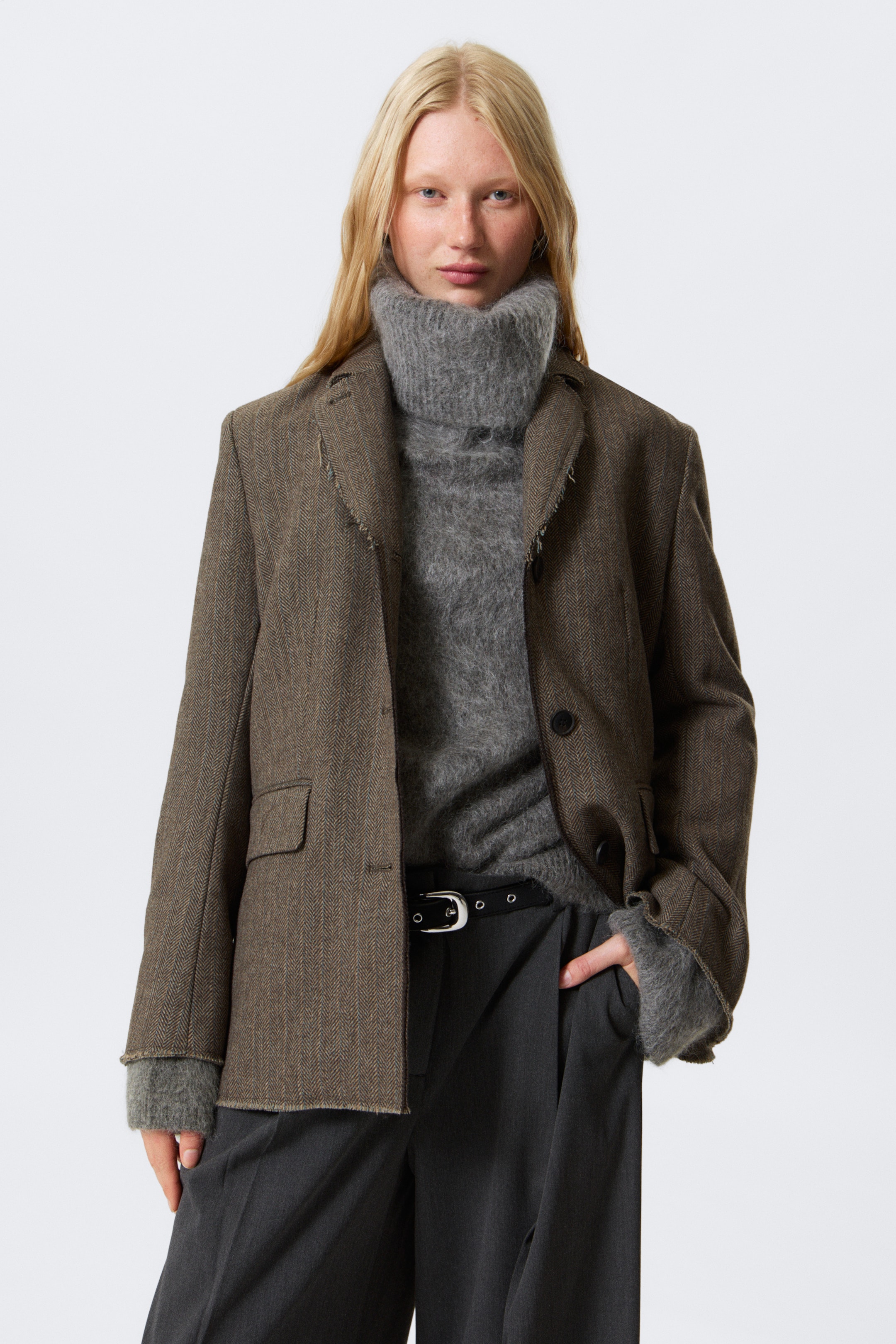 Vis større bilde: En brun blazer i fiskebensmønster bæres, vinklet litt til høyre, med frynsete kanter på slagene og falden. Den har en avslappet passform over en grå turtleneck-genser, hvis lange mansjetter strekker seg forbi blazerens ermer.