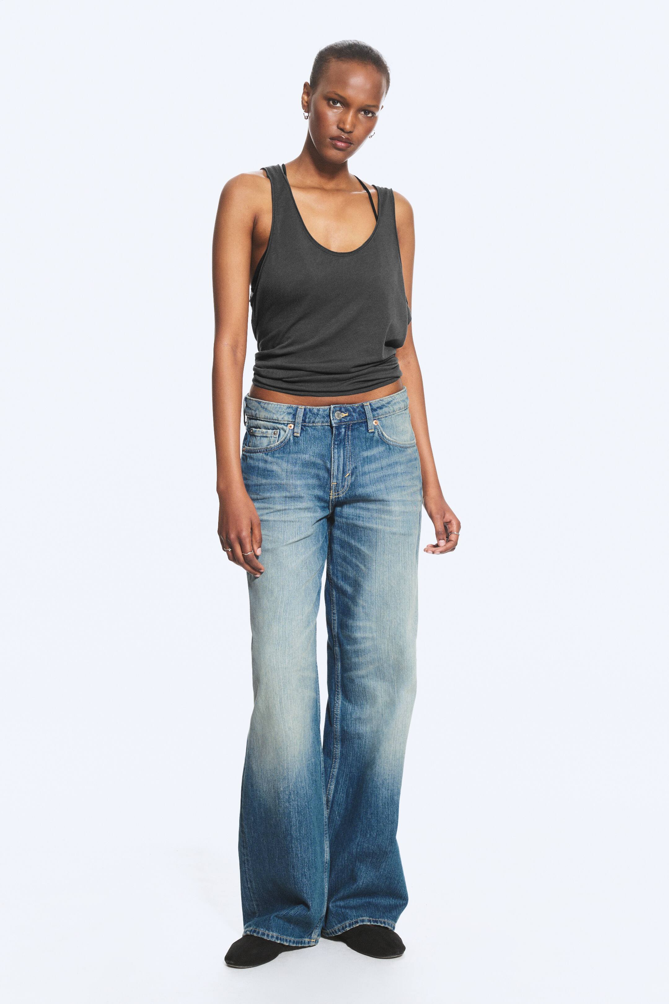 Vis større bilde: Lavt liv jeans med løs passform, vide ben og lang lengde bæres av modellen, som også har på seg en mørk tanktop som fremhever jeansens lave liv. Jeansene har en lys til medium blå vask med falming, et 5-lommers design og en synlig glidelås, med den lange lengden som dekker modellens sko.