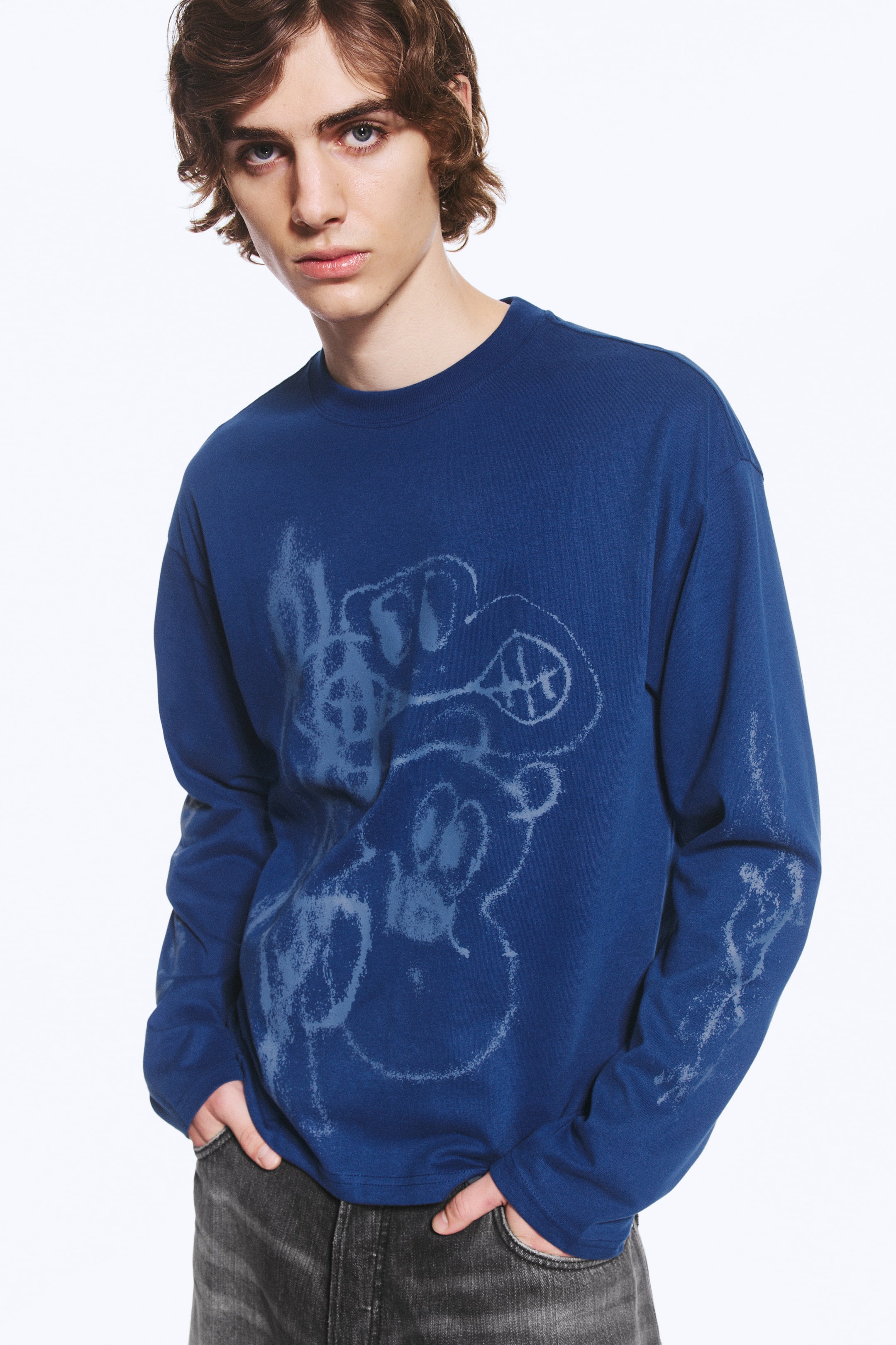 Vezi imaginea mai mare: Un tricou albastru, cu croială boxy și mâneci lungi, având un imprimeu grafic bleu pe partea din față și pe mânecă, este purtat de model. Modelul stă cu mâinile în buzunarele blugilor gri, evidențiind croiala lejeră a tricoului și gulerul rotund ribat.