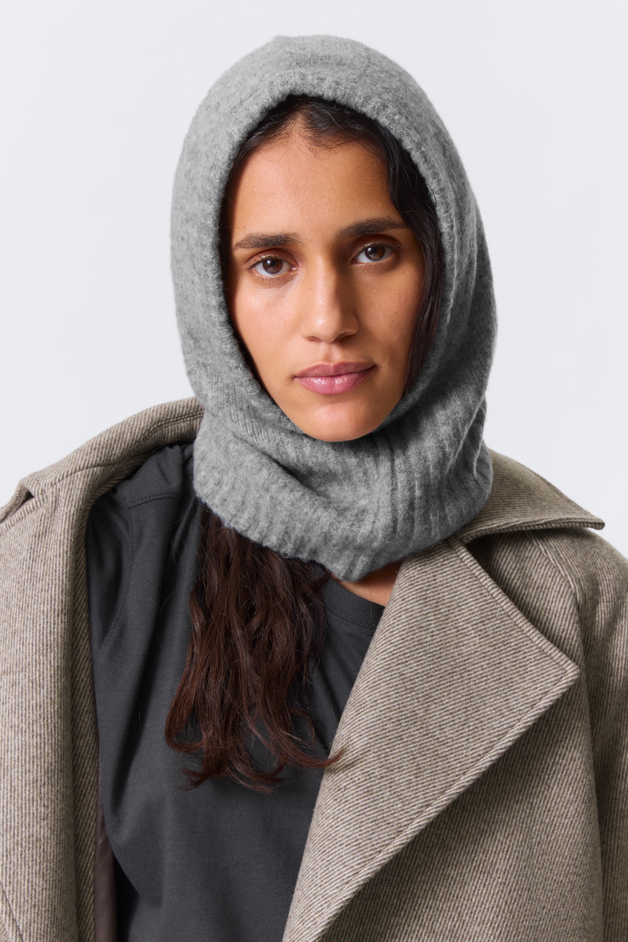 Agrandir l'image: Une cagoule grise en tricot de style balaclava s'ajuste parfaitement sur la tête et le cou d'une personne, présentée de face pour montrer son ajustement. La partie supérieure de la capuche présente un tricot lisse, tandis que la zone inférieure du cou arbore une texture côtelée. Elle est portée par-dessus un haut sombre et un manteau brun clair, démontrant son utilisation en superposition.