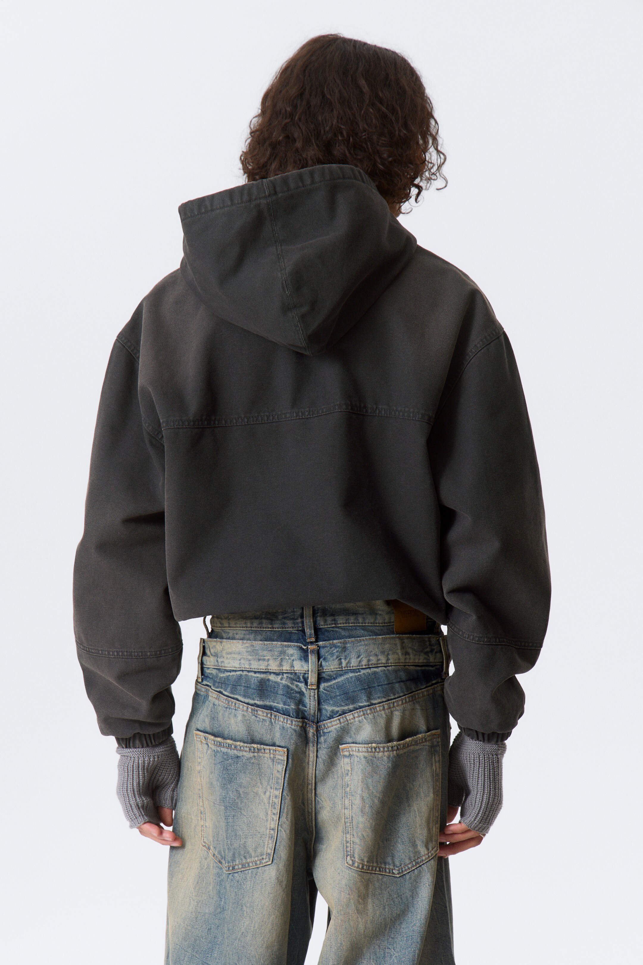 Vis større bilde: En mørk grå, vasket-effekt hettegenser presenteres fra baksiden, med hette, fremtredende sømdetaljer over øvre rygg, og vide ermer med sydde mansjetter. Lyseblå, distressed vide jeans med baklommer og grå ribbestrikkede fingerløse hansker bæres sammen med hettegenseren.