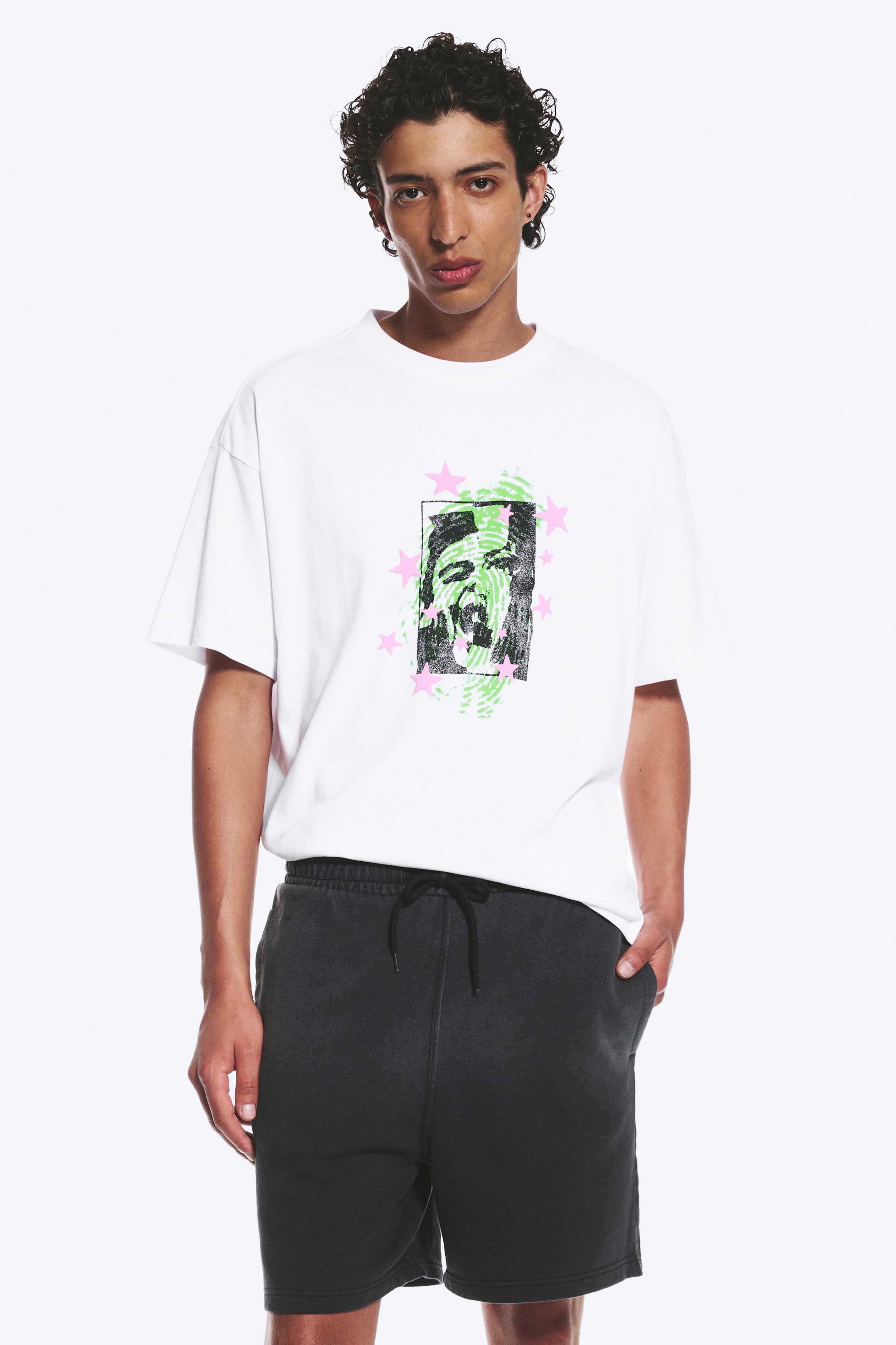 Visualizza immagine più grande: Una t-shirt bianca a maniche corte presenta una stampa grafica centrata di un volto astratto con texture verdi e stelle rosa all'interno di un contorno rettangolare nero. Dei pantaloncini sportivi grigio scuro completano il look, mostrando una cintura elasticizzata con un cordino nero.
