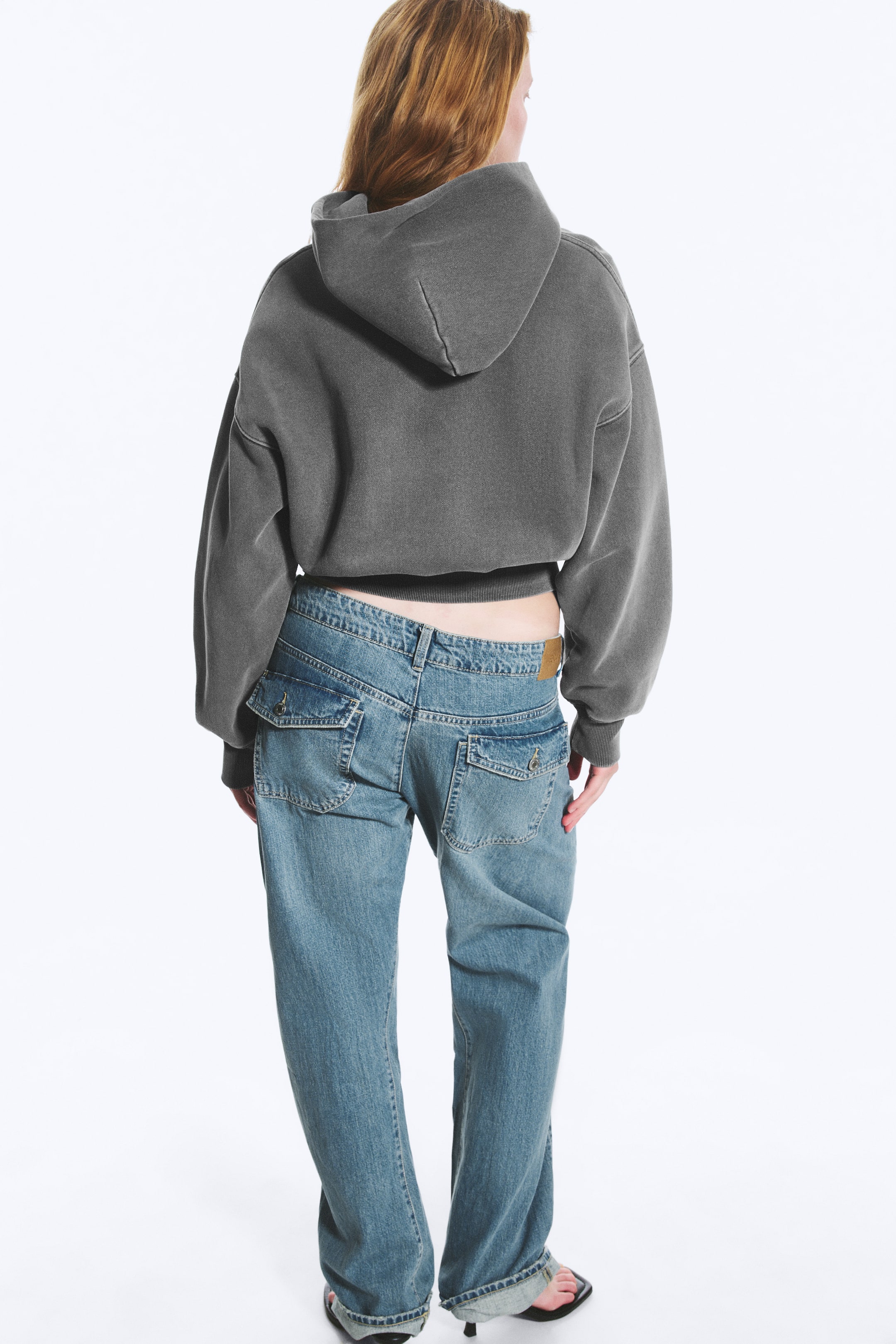Größeres Bild ansehen: Ein dunkelgrauer, verkürzter Hoodie ist von hinten zu sehen und zeichnet sich durch eine übergroße Passform mit gerippten Bündchen und einem gerippten Saum aus. Er wird mit hellblauen, tief sitzenden, weit geschnittenen Jeans kombiniert, die auf der Rückseite und an den Hüften Klappentaschen, einen braunen Lederbundaufnäher und hochgekrempelte Säume aufweisen.