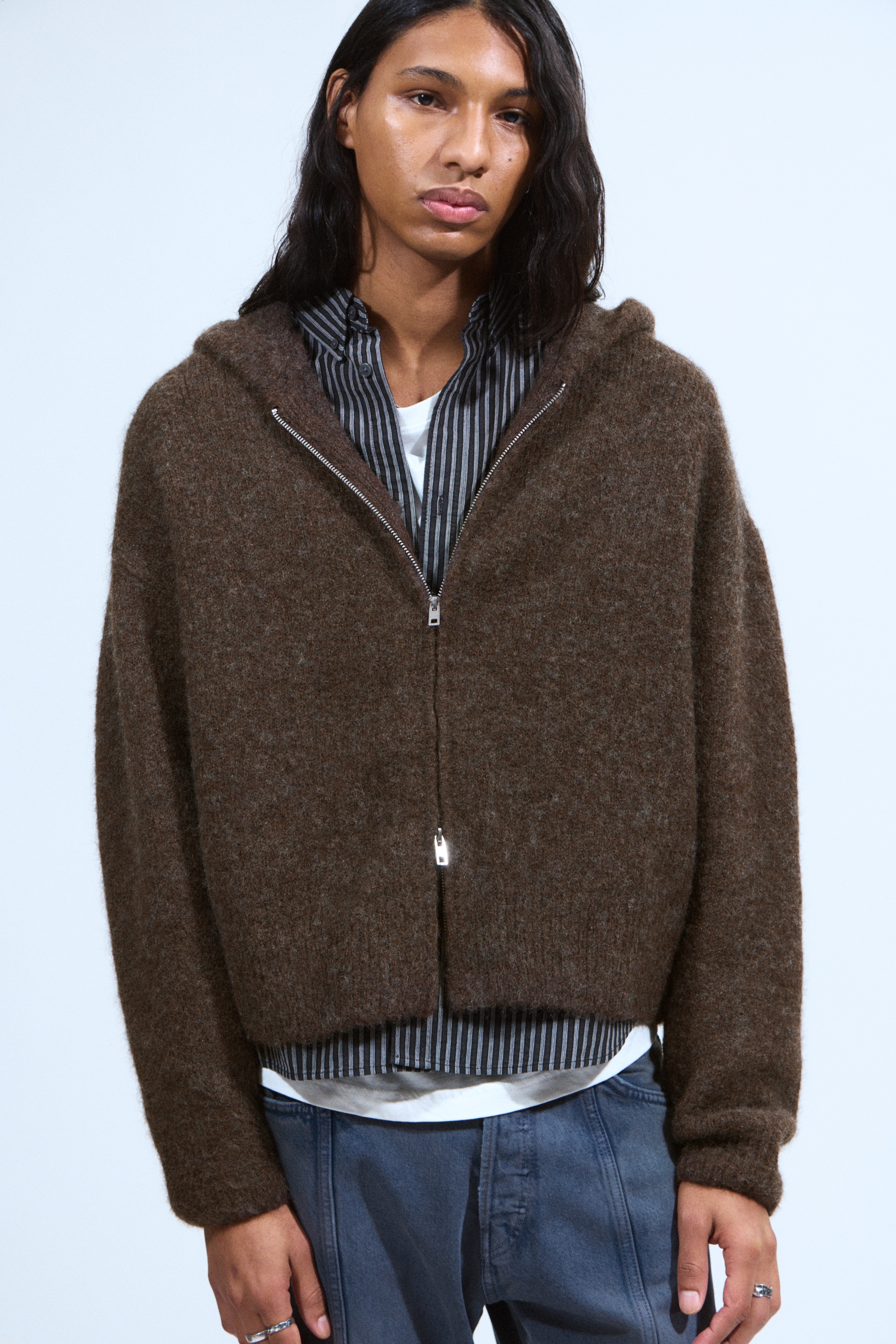 View larger image: A brown textured knit hooded cardigan is being worn, facing forward, featuring an oversized fit and a silver full-zip, two-way zipper. It is layered over a black and white striped long-sleeved shirt and a white t-shirt.