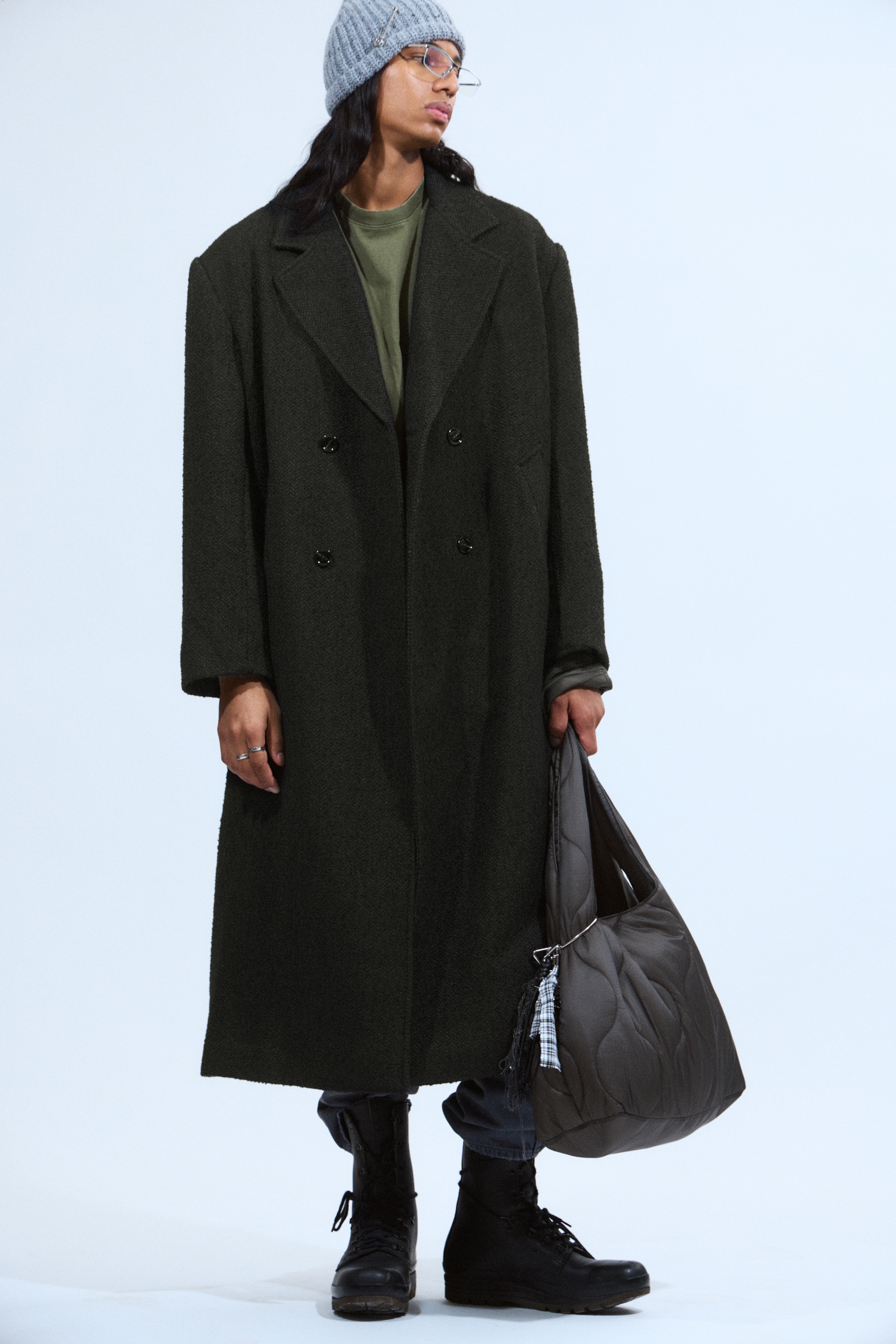 Ingrandisci l'immagine: Un cappotto doppiopetto verde scuro viene mostrato, caratterizzato da ampi revers a lancia e un tessuto strutturato. Il lungo cappotto, che si estende sotto il ginocchio, presenta una vestibilità oversize con spalle scese e due tasche con patta visibili, indossato sopra una t-shirt verde più chiara.
