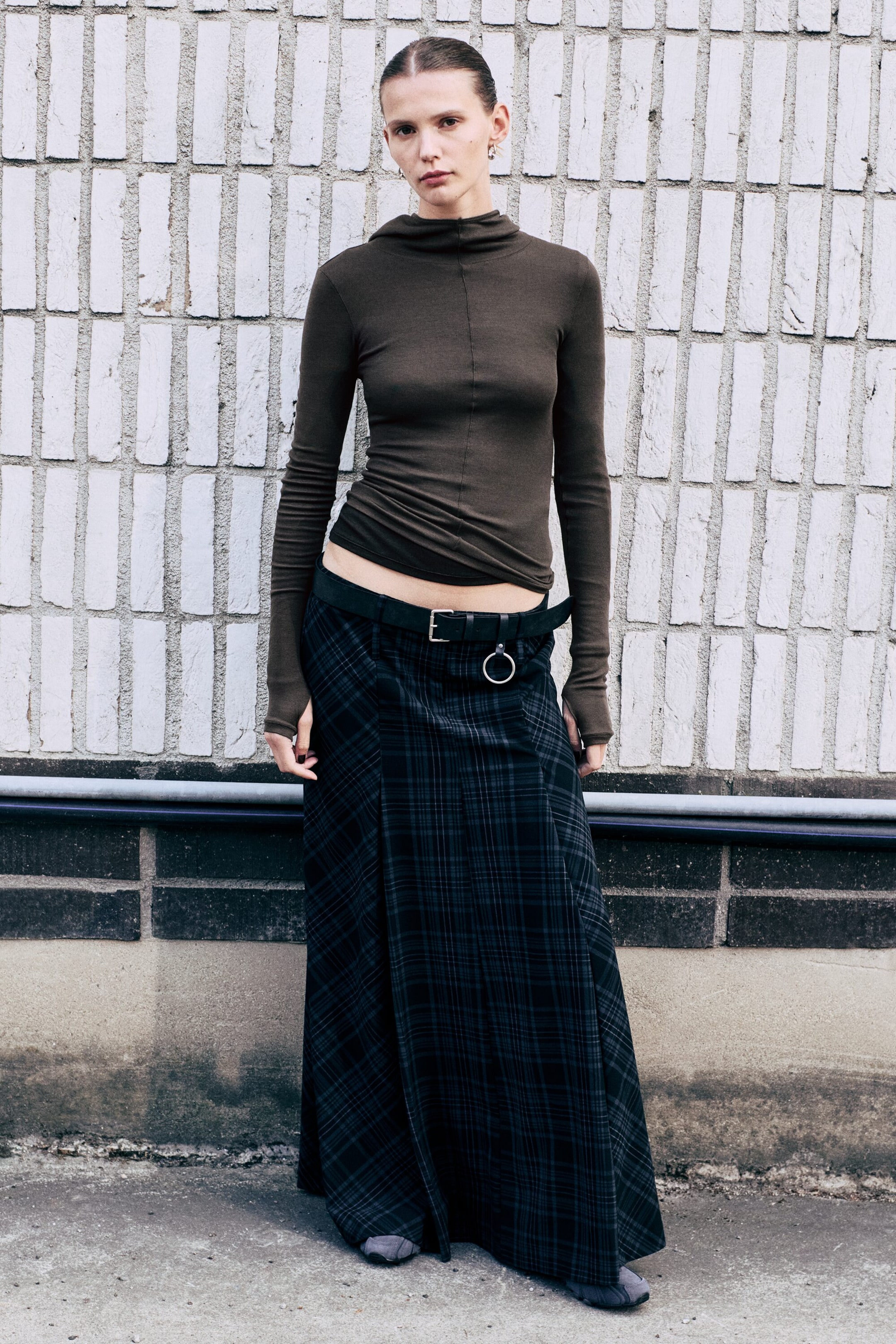 Größeres Bild ansehen: Ein Maxirock mit niedriger Taille, dunkel kariertem Muster und ausgestellter Silhouette mit Panel-Konstruktion wird von dem Model getragen. Der Rock ist mit einem schwarzen Gürtel mit silberfarbenem Ring kombiniert und zusammen mit einem figurbetonten Langarm-Mock-Neck-Top gestylt.