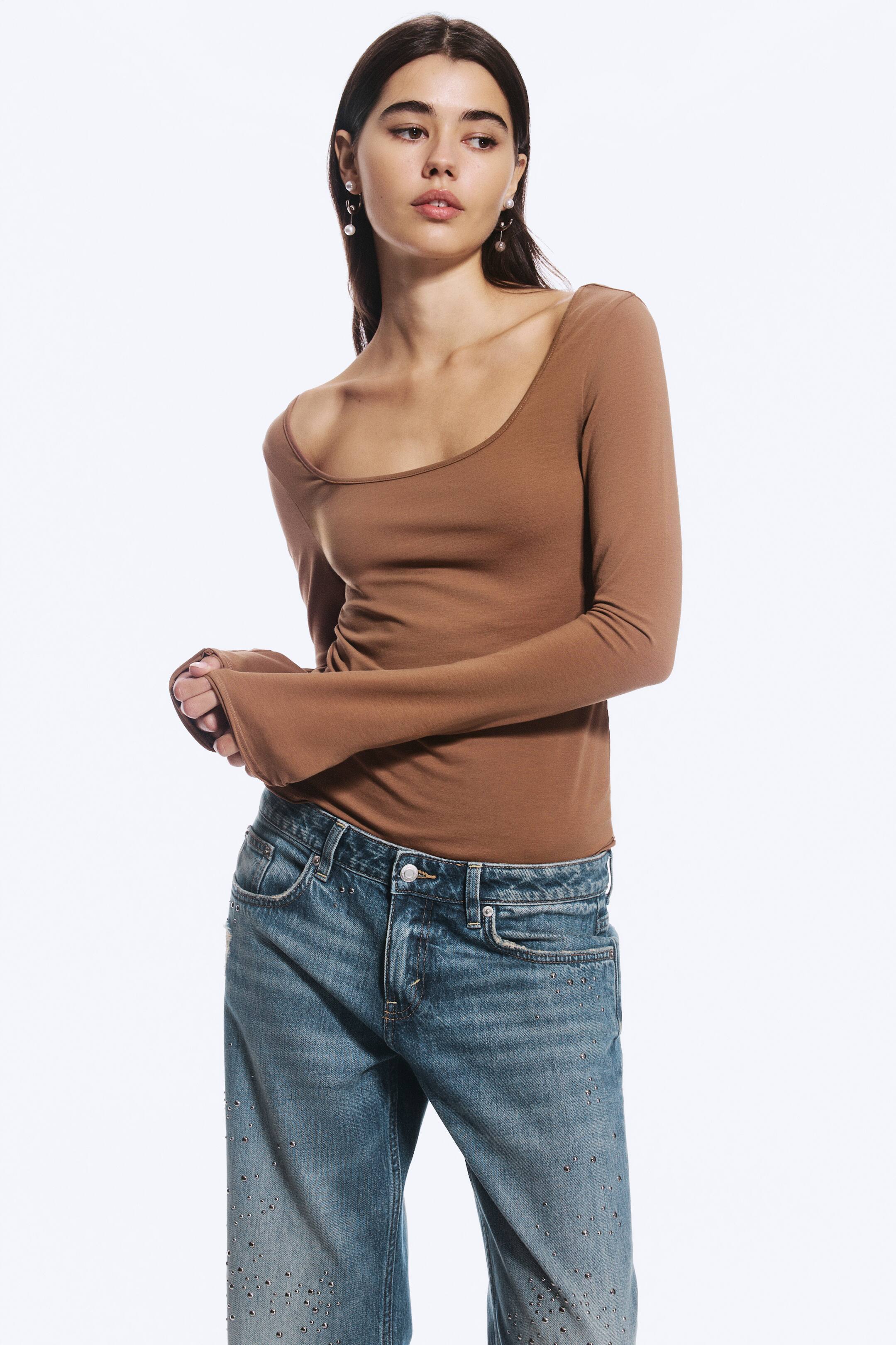 Größeres Bild ansehen: Ein braunes Oberteil mit langen Ärmeln, weitem Rundhalsausschnitt und schmaler Silhouette wird vom Model getragen und mit verzierten blauen Jeans kombiniert. Das Model hält die verlängerten Ärmelbündchen und setzt so das eng anliegende Design des Oberteils in Szene.
