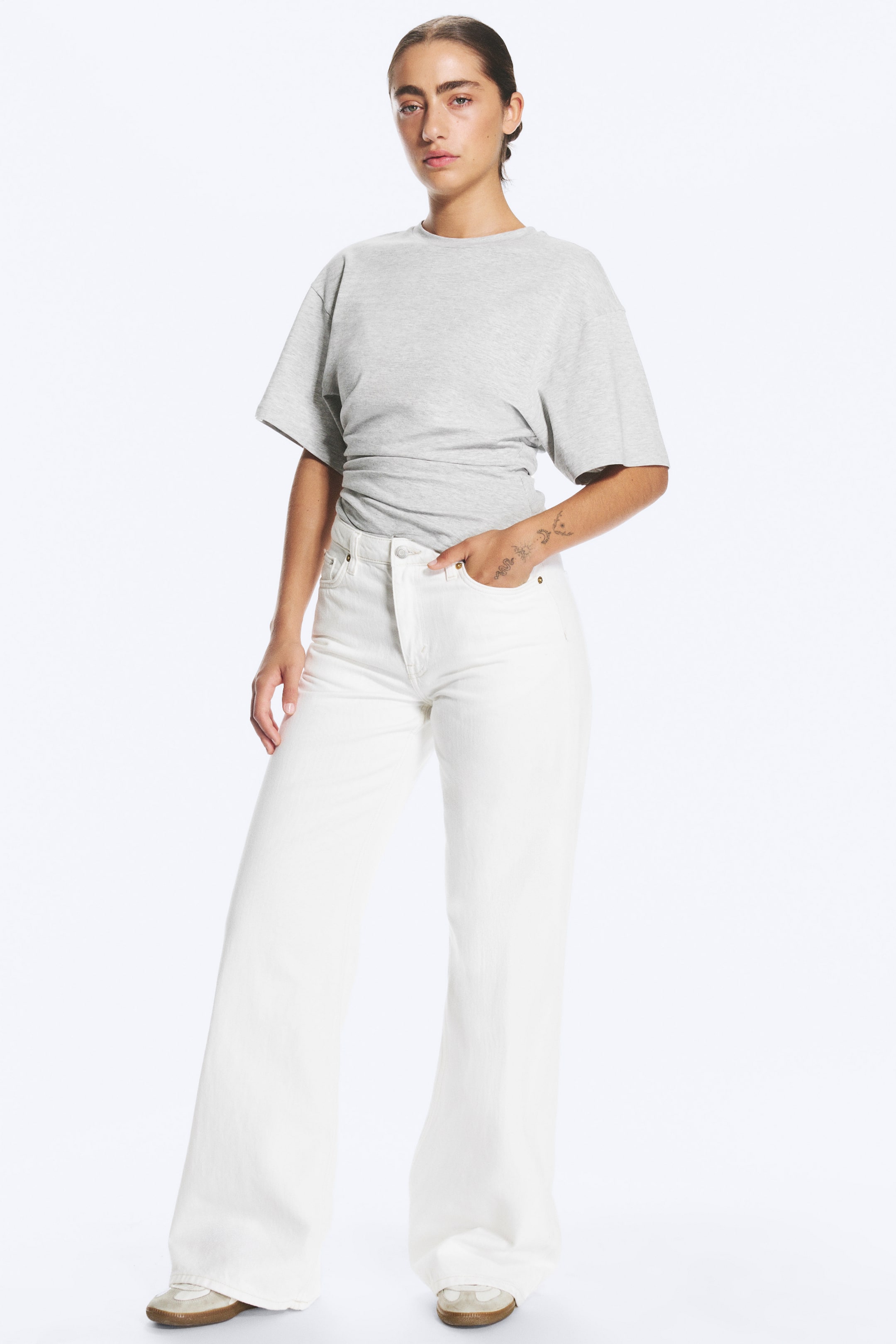 Größeres Bild ansehen: Die weiße Mid-Rise-Jeans mit weitem Bein wird vom Model getragen, das mit einer Hand in der vorderen Tasche steht. Dazu kombiniert ist ein in die Hose gestecktes graues T-Shirt mit kurzen Ärmeln.