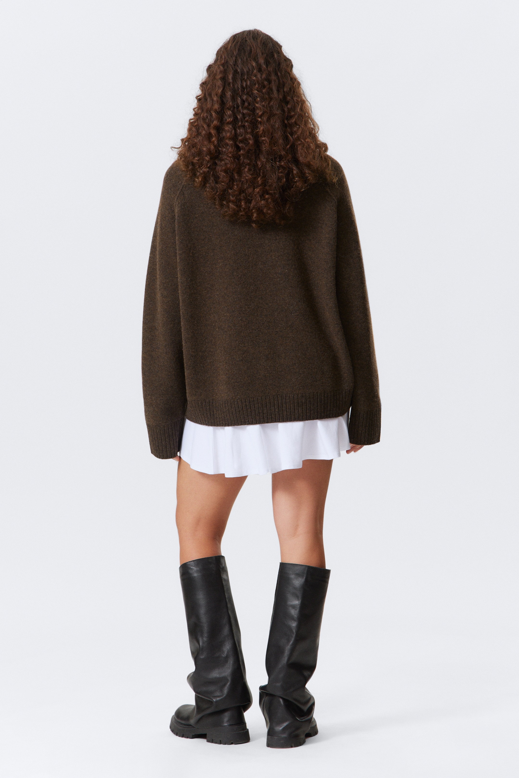 Größeres Bild anzeigen: Ein übergroßer, dunkelbrauner Strickpullover wird von hinten gezeigt, mit einer lockeren Passform, langen Ärmeln und einem gerippten Saum. Ein weißes Hemd wird darunter geschichtet, dessen plissierter Saum unter dem Pullover sichtbar ist.