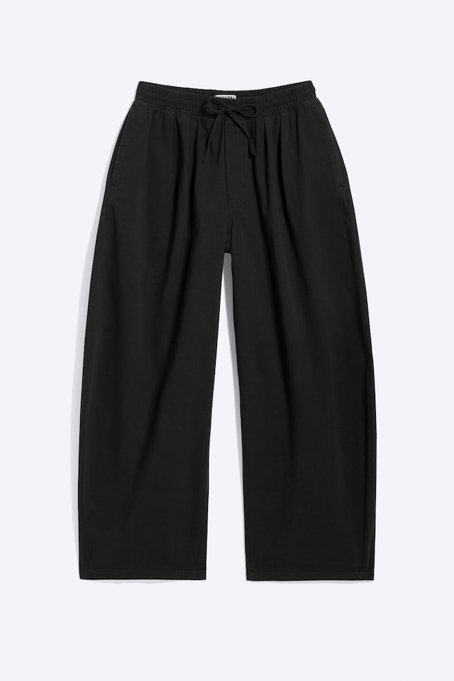 Pantalones negros vistos desde el frente, con una cintura elástica fruncida y un cordón ajustable. Se aprecian bolsillos en las costuras laterales, y las piernas anchas terminan en un dobladillo recto. - 1