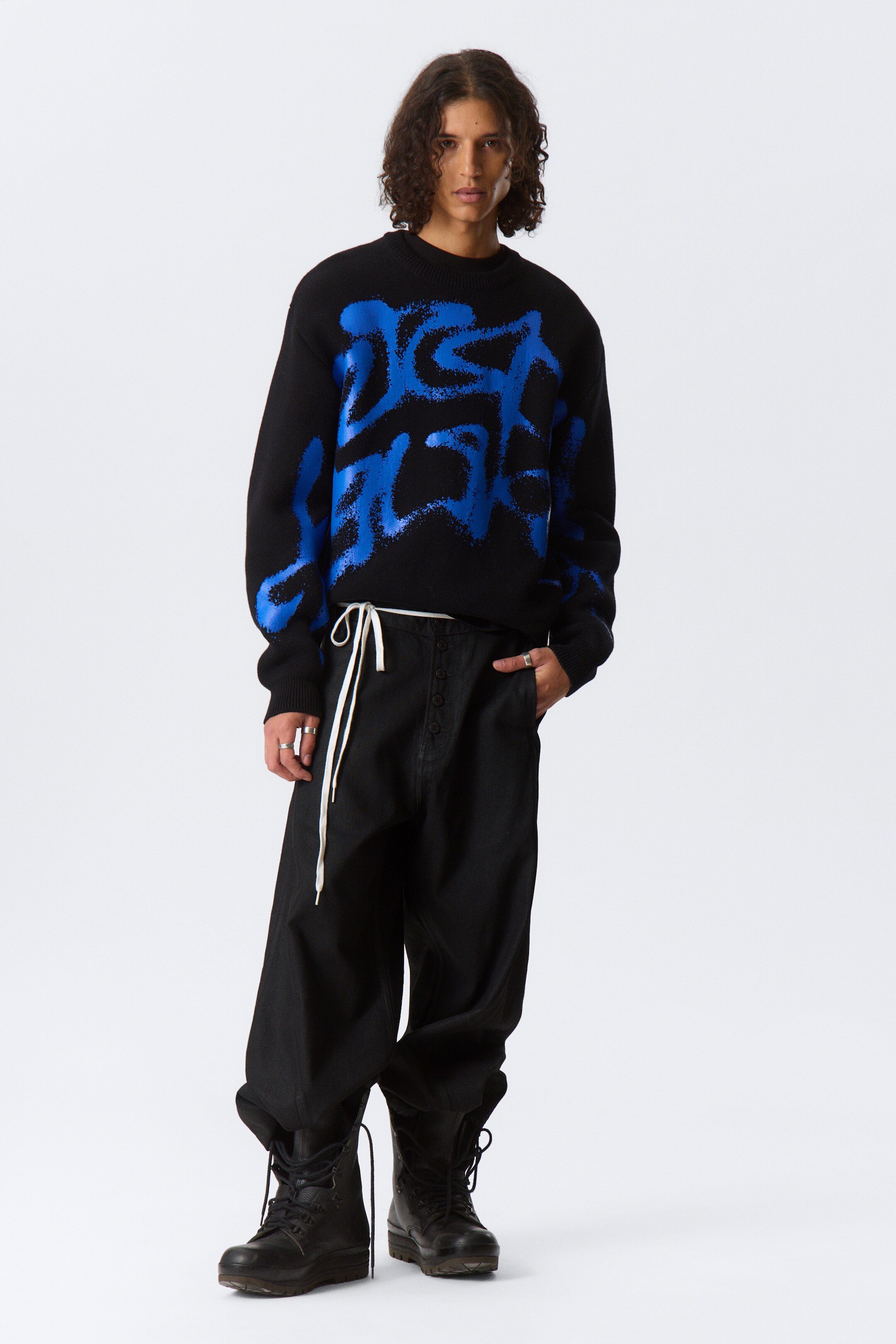 Se større billede: En sort jacquard-strikket grafisk sweater med blå mønstre bæres af modellen. Den har en rund ribkantet halsudskæring, ribbede manchetter og en ribkantet søm.