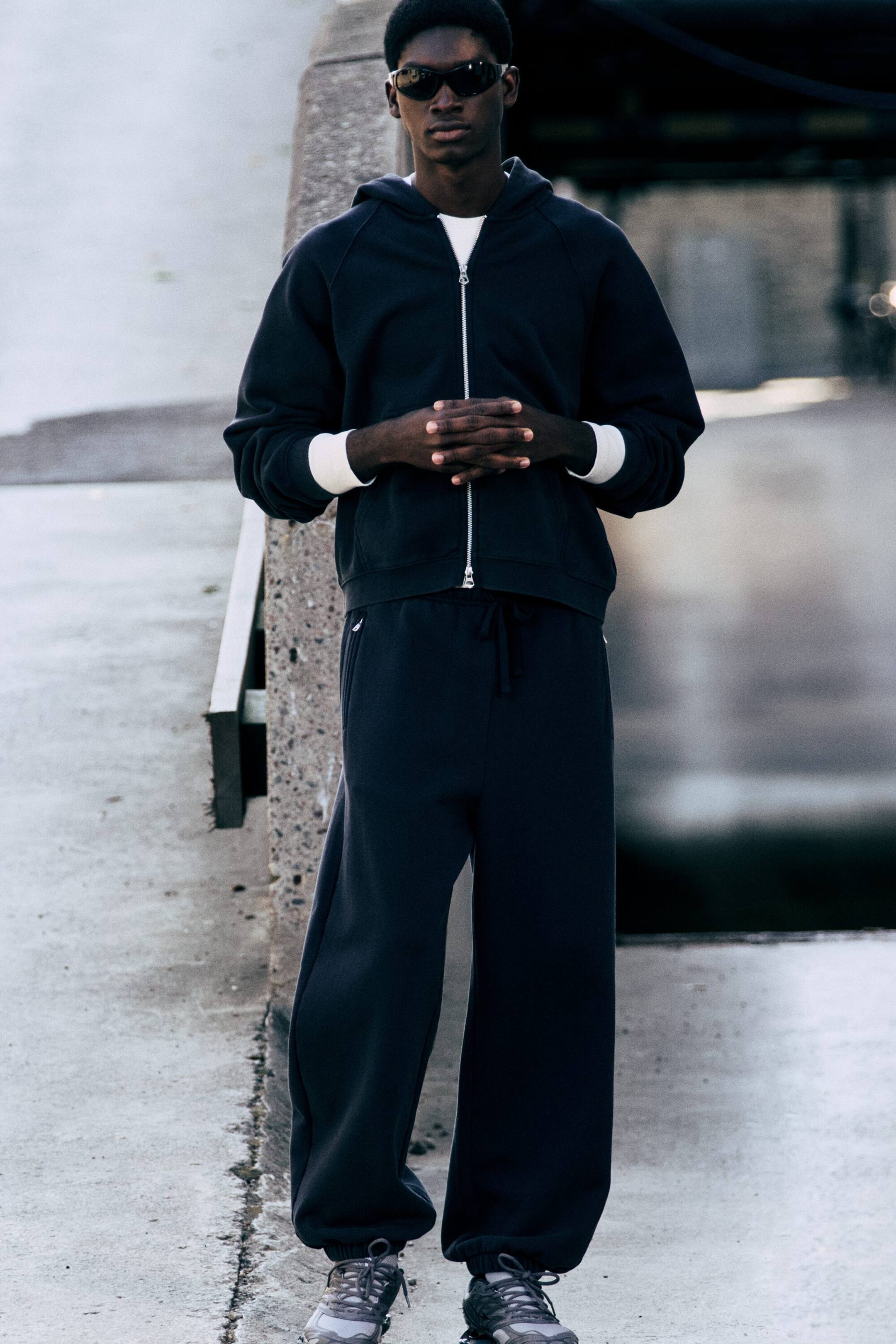 Visualizza l'immagine ingrandita: Il modello indossa pantaloni della tuta grigio scuro dalla vestibilità ampia e con gamba a barilotto, dotati di vita elasticizzata con coulisse piatta e tasche a filetto con zip. I pantaloni sono rifiniti con polsini elasticizzati alle caviglie e abbinati a una felpa con cappuccio con zip e sneaker coordinate.