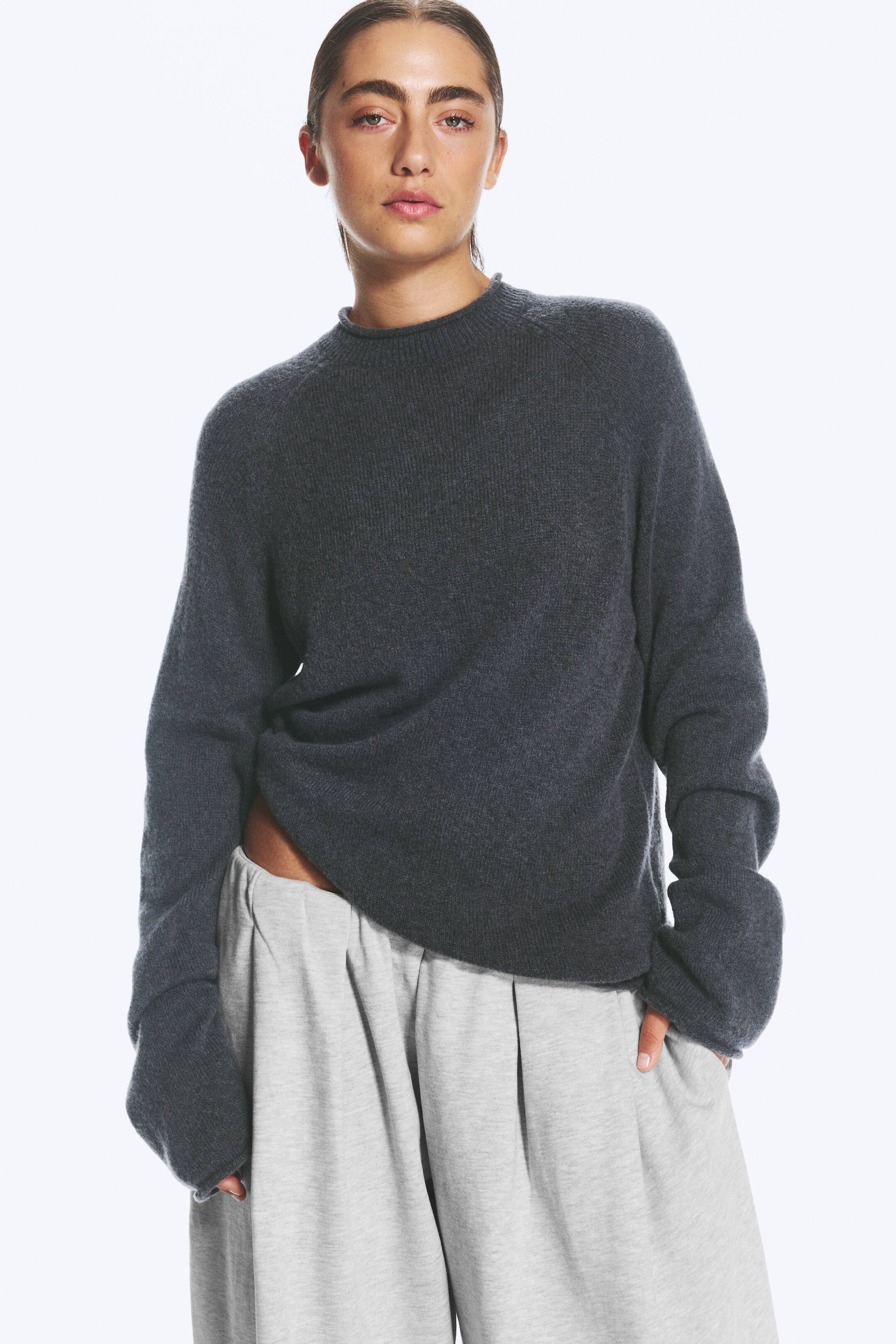 Visualizza immagine più grande: Un maglione lavorato a maglia grigio scuro, dalla vestibilità morbida e con maniche lunghe, indossato dalla modella. Il maglione presenta un alto colletto a imbuto con bordo a taglio vivo ed è abbinato a pantaloni larghi grigio chiaro.