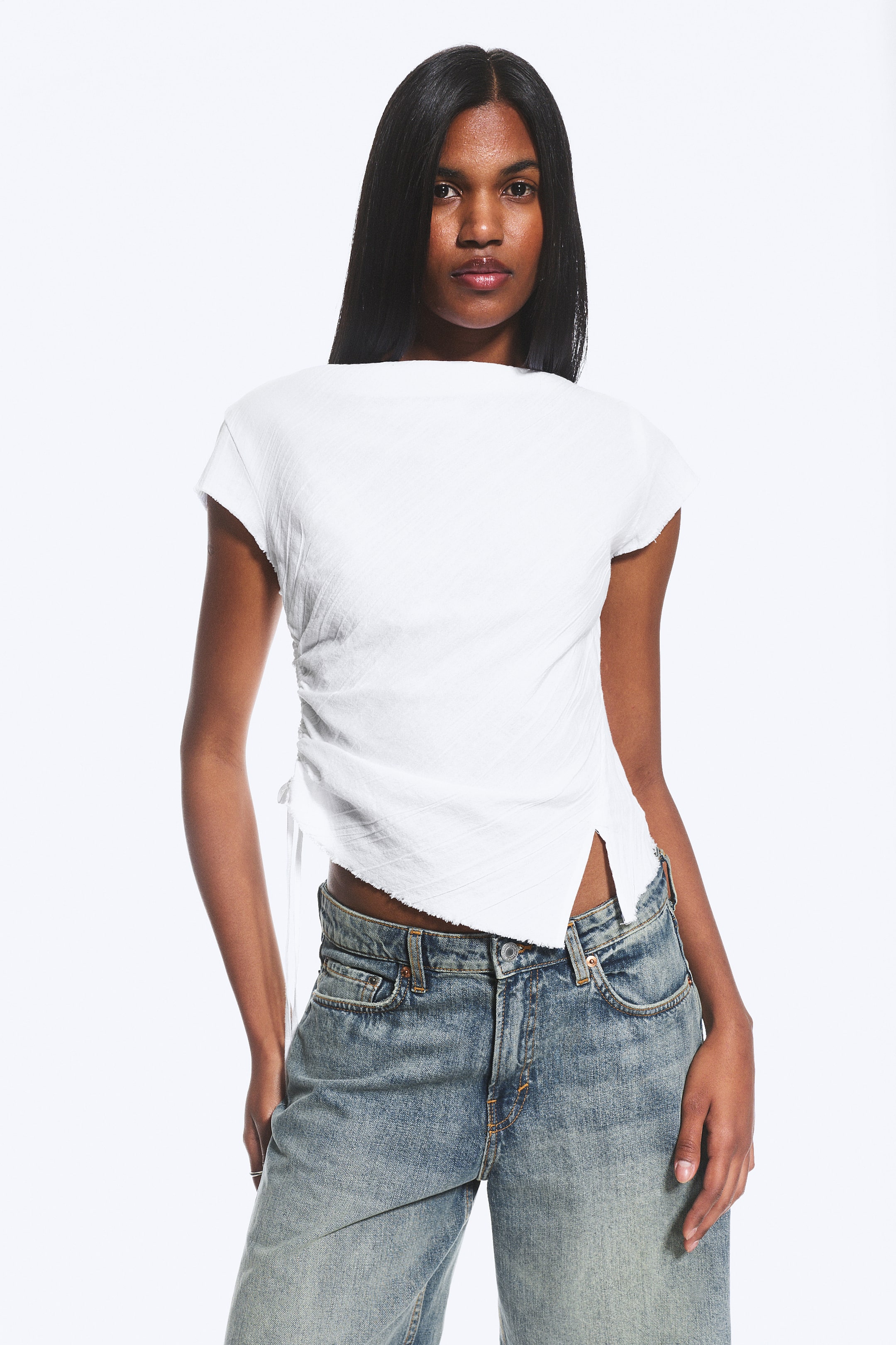 View larger image: A white textured short-sleeved top is showcased, featuring a wide neckline and ruched detailing with ties on the left side. Its asymmetrical raw-edged hemline includes a slit on the right, resting over blue jeans.
