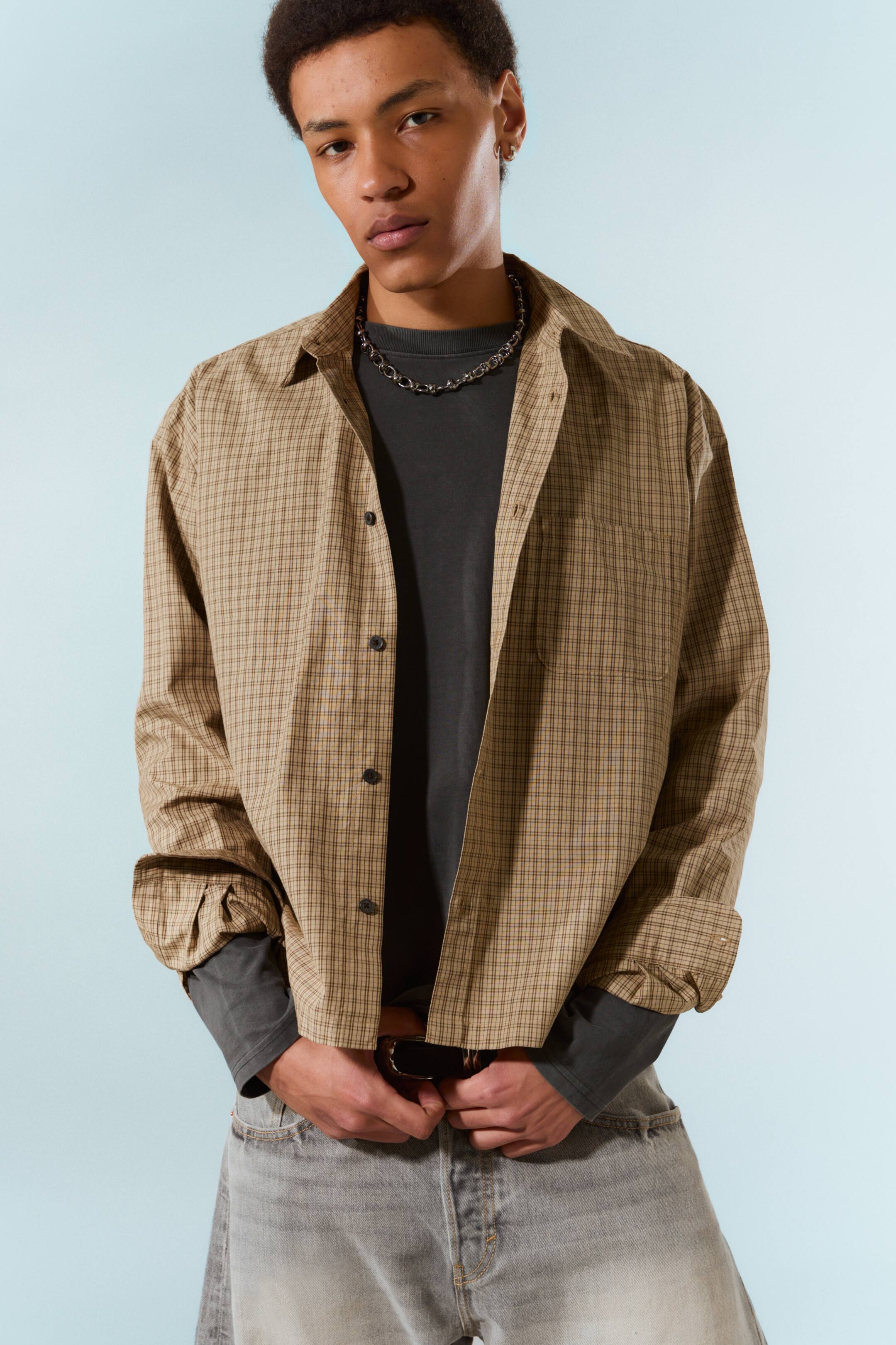 Grotere afbeelding bekijken: Een bruine en beige geruite overhemd met een gespreide kraag, donkere knopen aan de voorkant en een borstzak. De lange mouwen zijn opgerold en het wordt open gedragen over een donkergrijs T-shirt met lange mouwen.