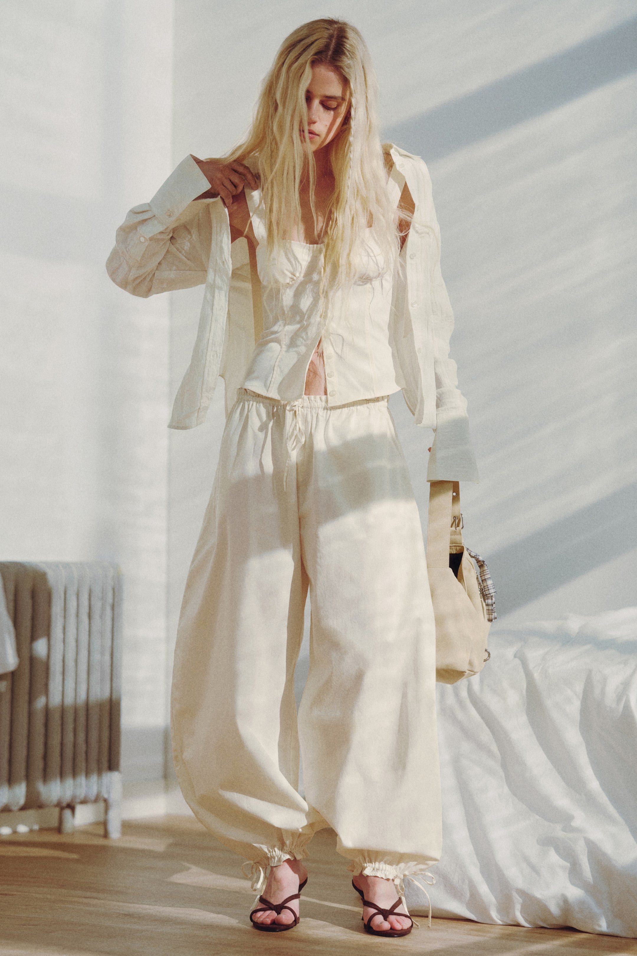 View larger image: An off-white ensemble features a long-sleeved shirt, worn open over a thin-strapped, button-front bustier-style top. Matching wide-leg trousers showcase a drawstring waist and gathered ankles with ties. Dark brown strappy sandals and a light beige shoulder bag accompany the outfit.