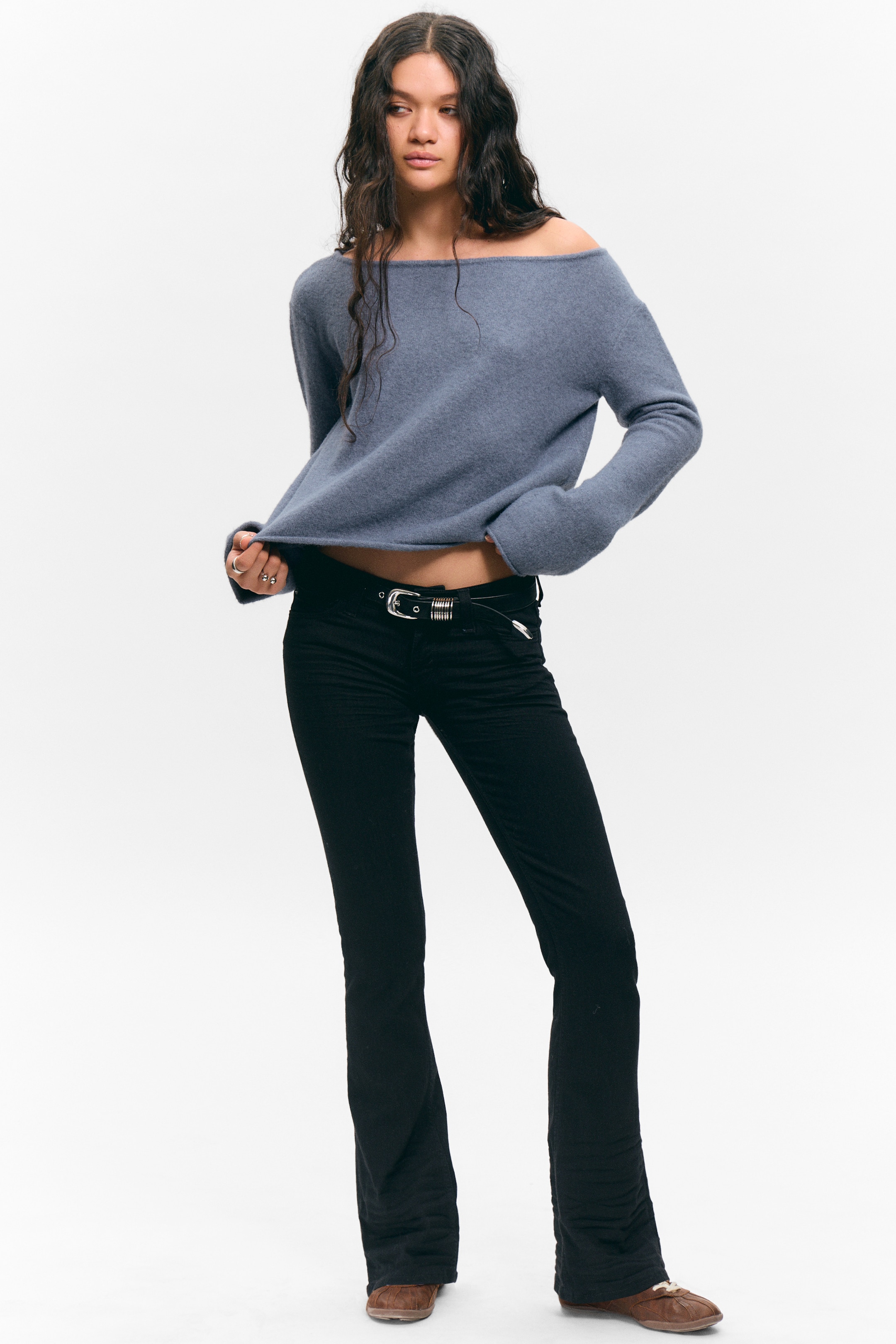 Größeres Bild ansehen: Ein gedämpfter blauer, schulterfreier Pullover wird präsentiert, der über einen breiten Bootsausschnitt und lange Ärmel verfügt. Er wird mit schwarzen, tief sitzenden Schlaghosen gestylt, die eine subtile gerippte Textur aufweisen und mit einem dünnen schwarzen Gürtel mit silberner Schnalle kombiniert sind, während braune flache Schuhe das Ensemble vervollständigen.