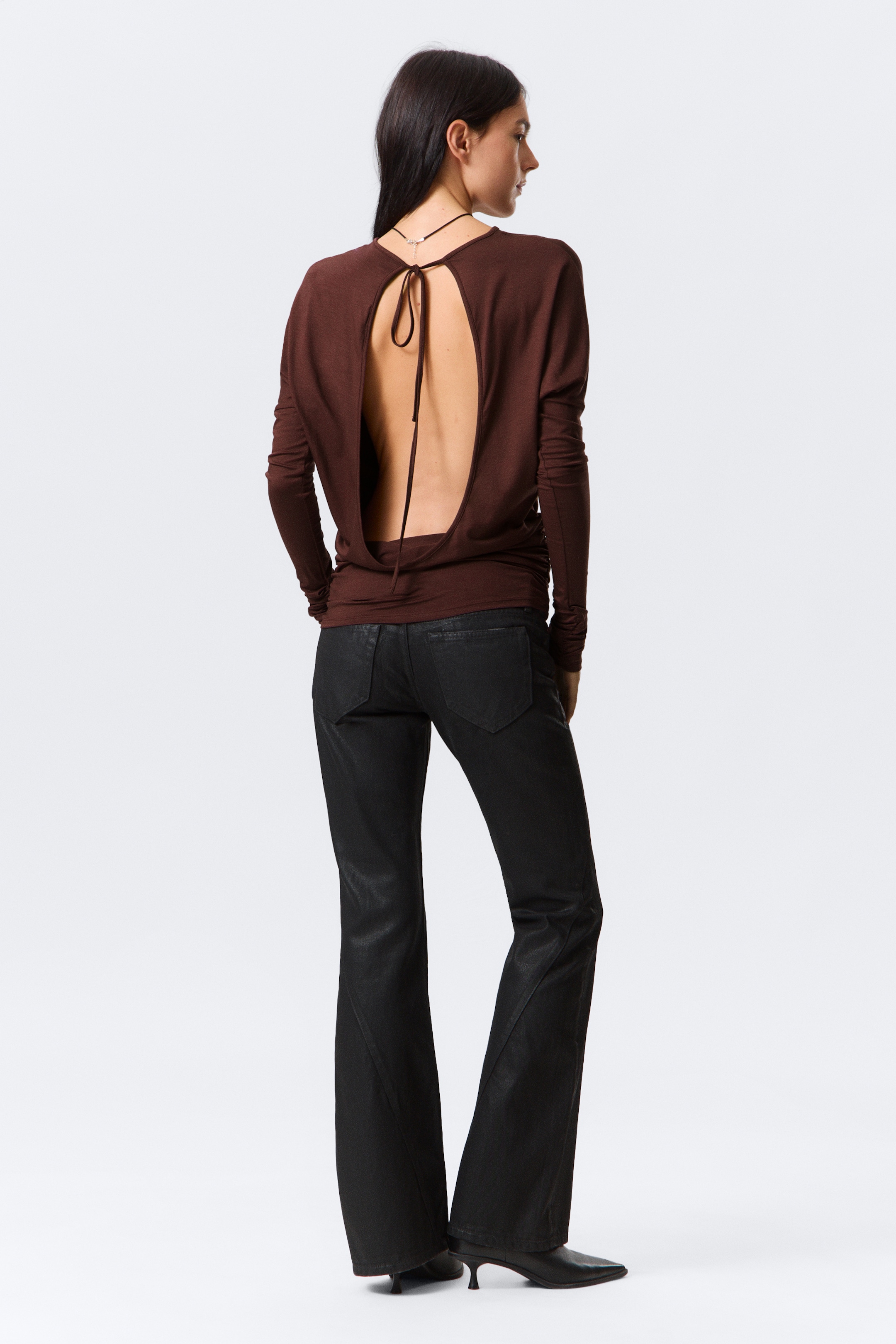 Agrandir l'image: Un haut à manches longues de couleur brun foncé est présenté de dos, mettant en valeur un grand dos ouvert de forme ovale, maintenu par de fines attaches au niveau du haut du dos. Il est assorti à un pantalon évasé noir subtilement enduit.