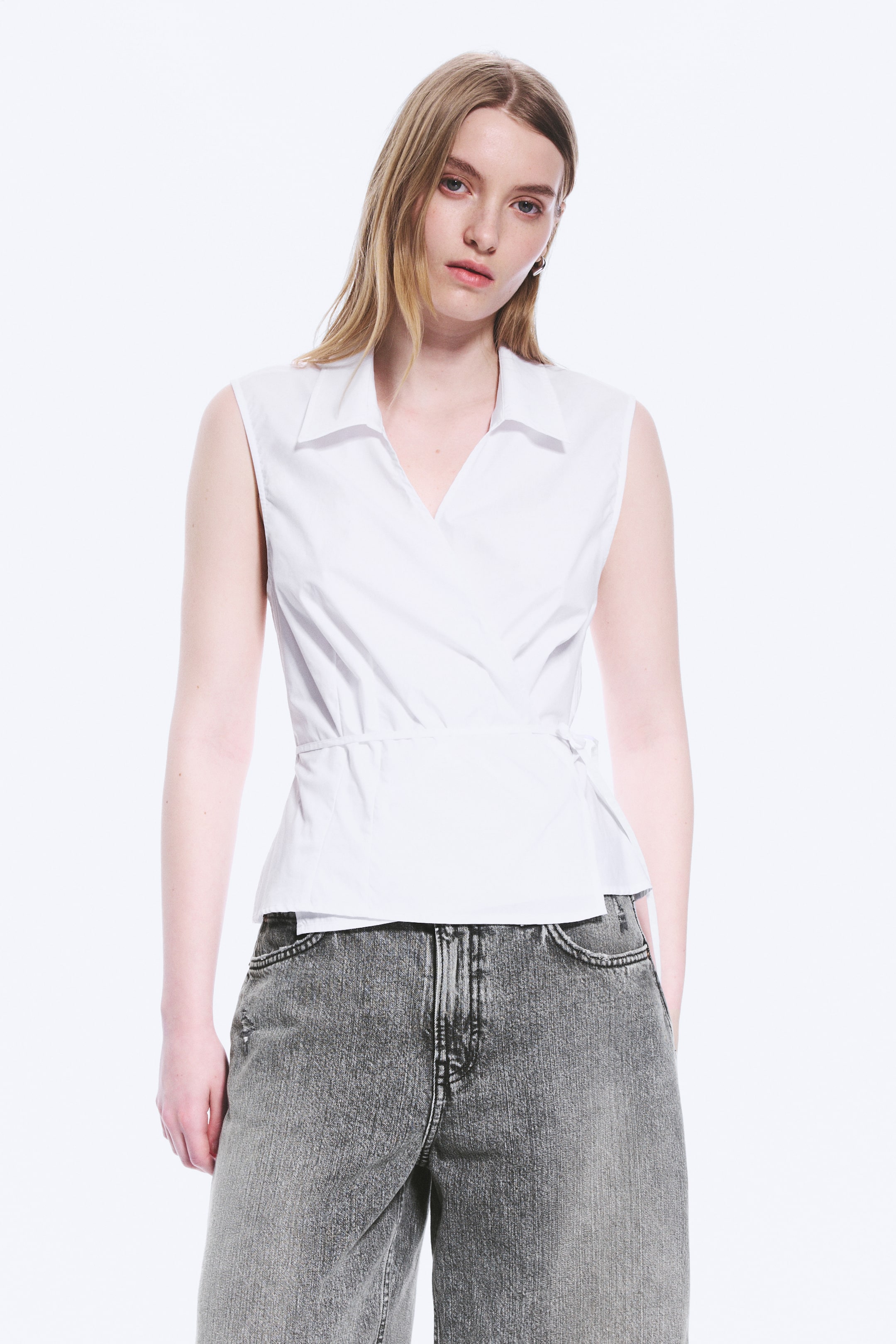 Visualizza immagine più grande: Un top bianco senza maniche con design a portafoglio presenta un colletto a punta e uno scollo a V, con il suo design avvolgente che si stringe in vita e si chiude con un laccio laterale visibile.