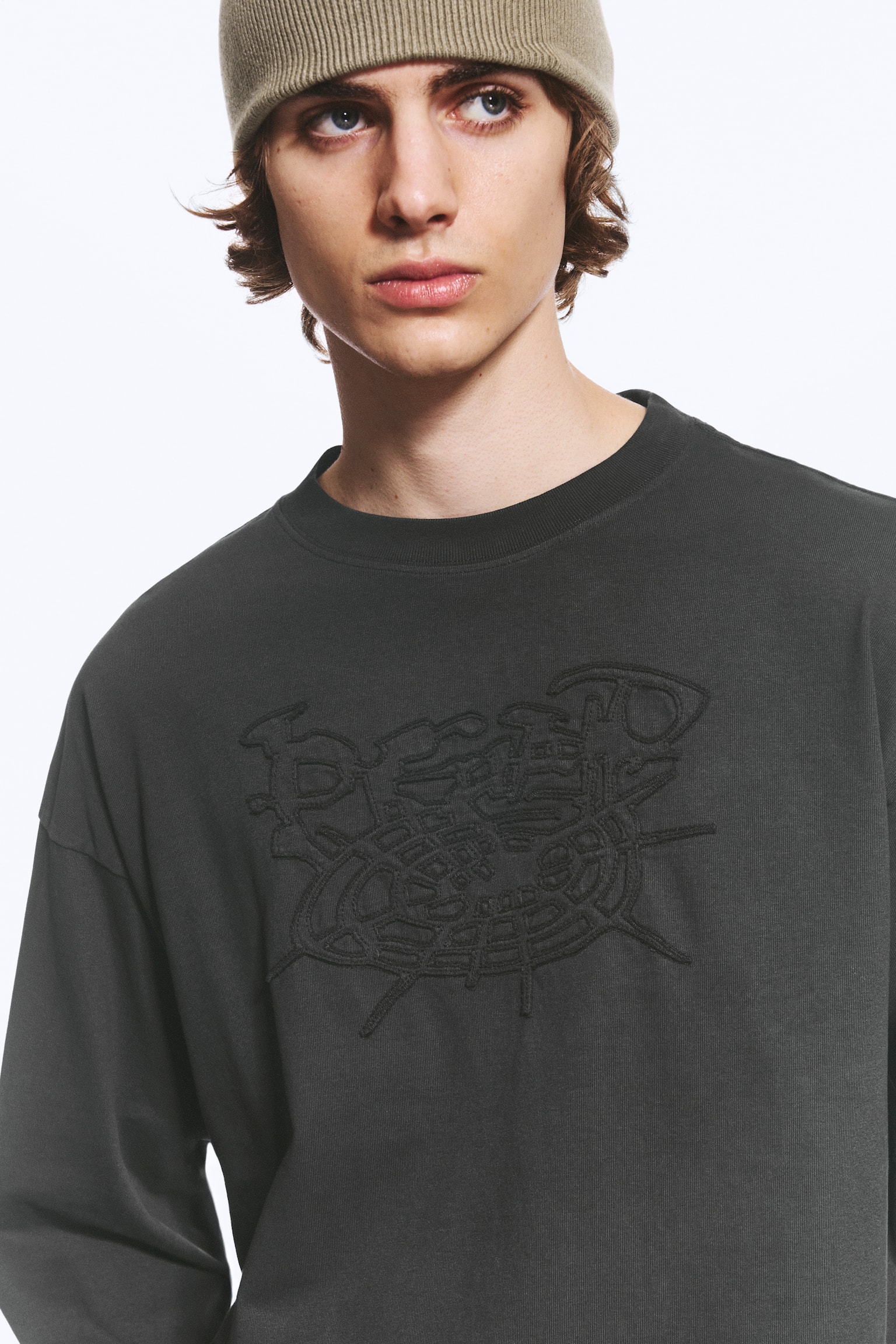 En mörkgrå boxig långärmad T-shirt visas framifrån och har en rund ribbad halsringning, nedhasade axlar och ett ton-i-ton-broderat motiv mitt på bröstet. - 1