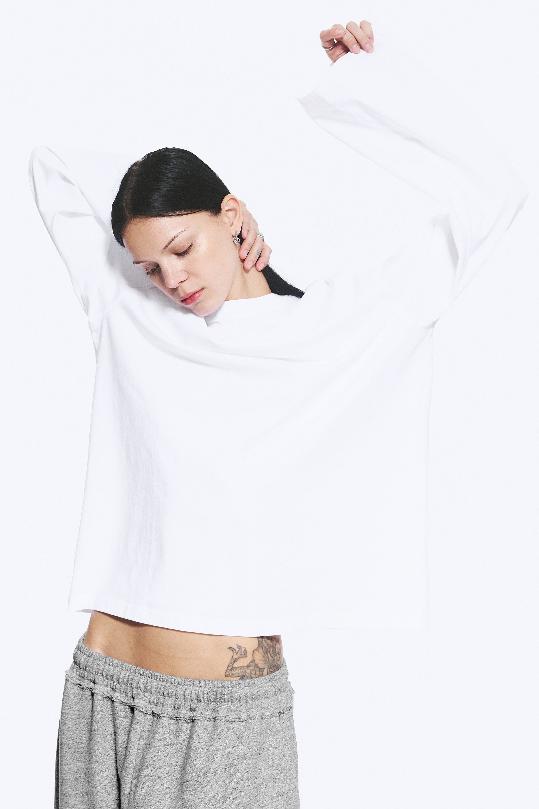 Se større billede: En hvid langærmet t-shirt bæres, med rund hals og en afslappet, oversized pasform. De lange ærmer strækker sig opad, og en kort, lige kant er synlig over taljen.
