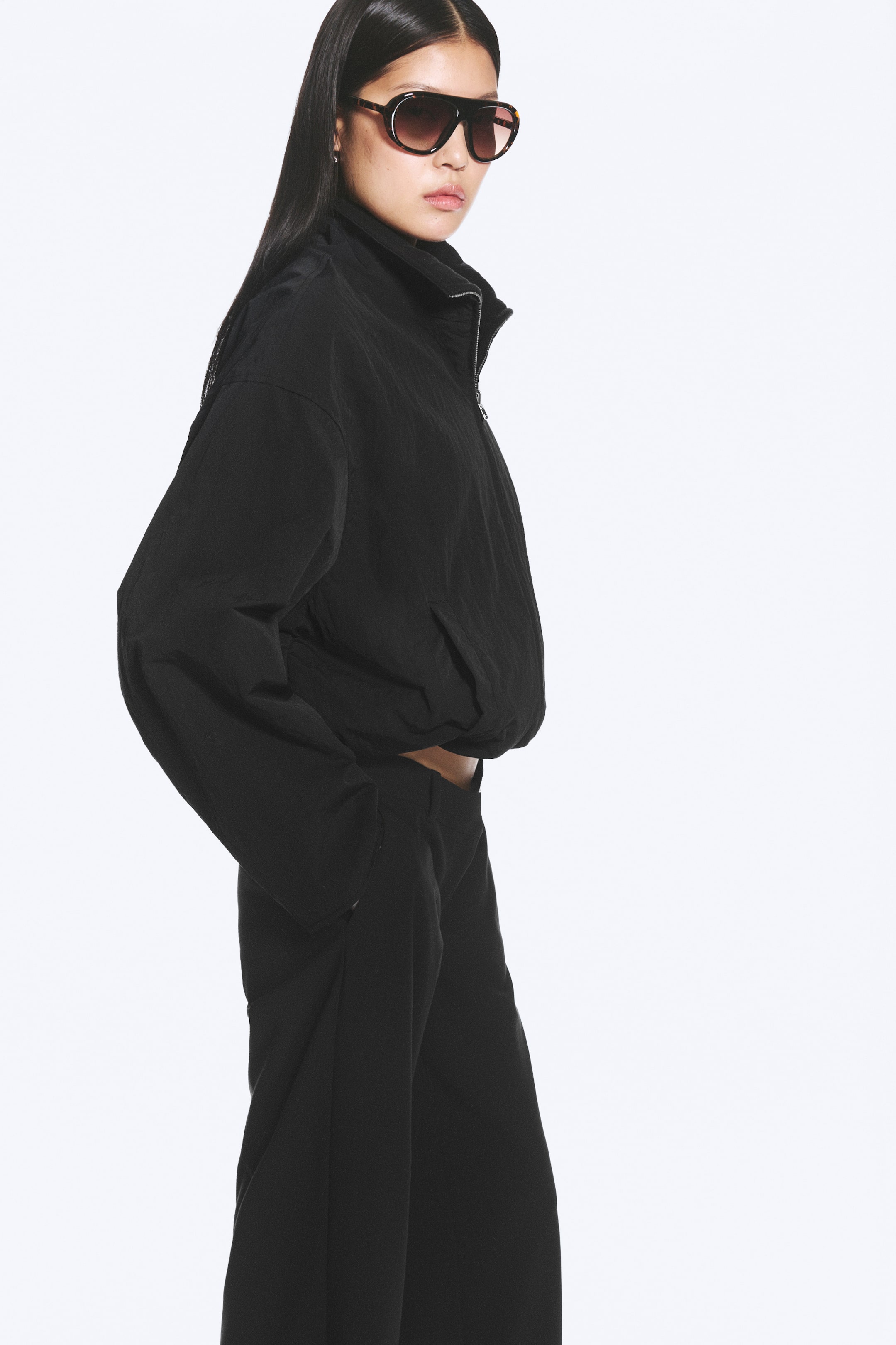 Visualizza immagine più grande: La modella indossa una giacca oversize nera. Presenta un colletto a imbuto, una chiusura frontale con zip parziale, maniche lunghe con spalle scese e un orlo arricciato. La modella ha una mano infilata in una tasca laterale.