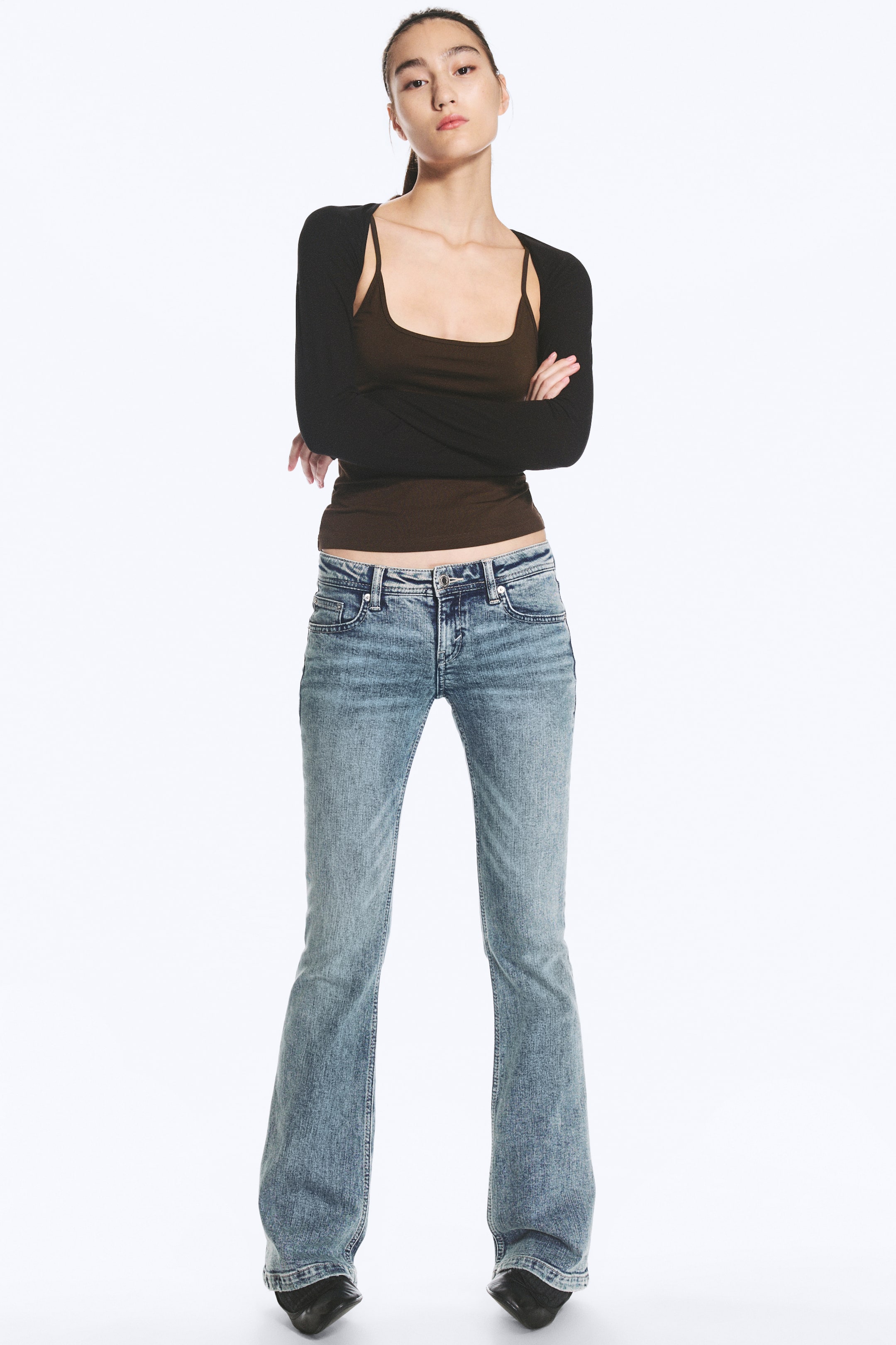 Visualizza immagine più grande: Un paio di jeans blu chiaro a vita bassa, dalla vestibilità slim e con gamba svasata, indossati dalla modella. I jeans presentano un classico design a cinque tasche con delicato effetto slavato e sono abbinati a una canottiera marrone e a un coprispalle nero a maniche lunghe.
