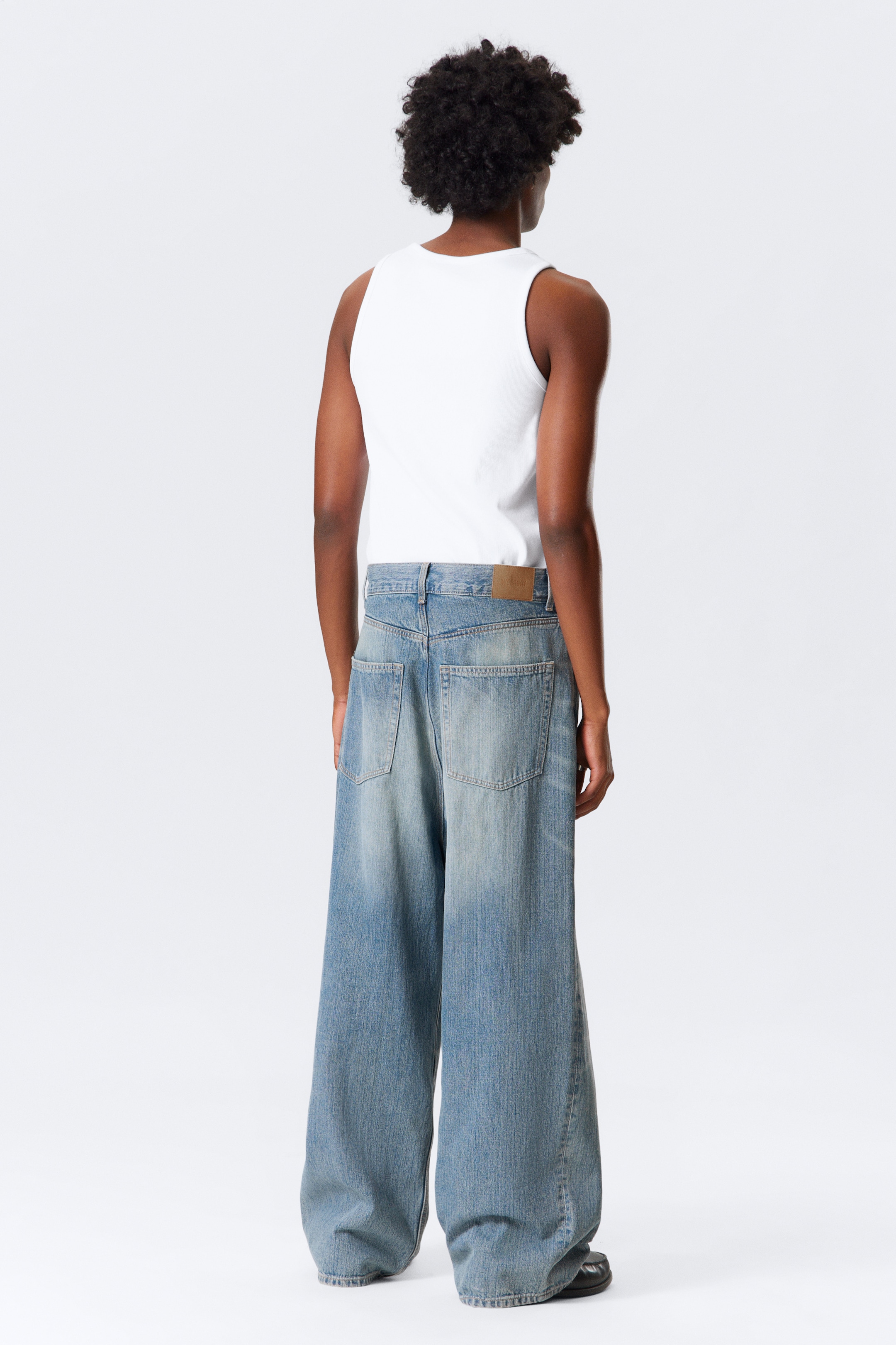 View larger image: Light blue, wide-leg jeans are being presented from the back, showcasing a baggy fit that is pooling at the ankles. The jeans are featuring two large back pockets and a brown patch on the rear waistband, and are being worn with a white tank top.