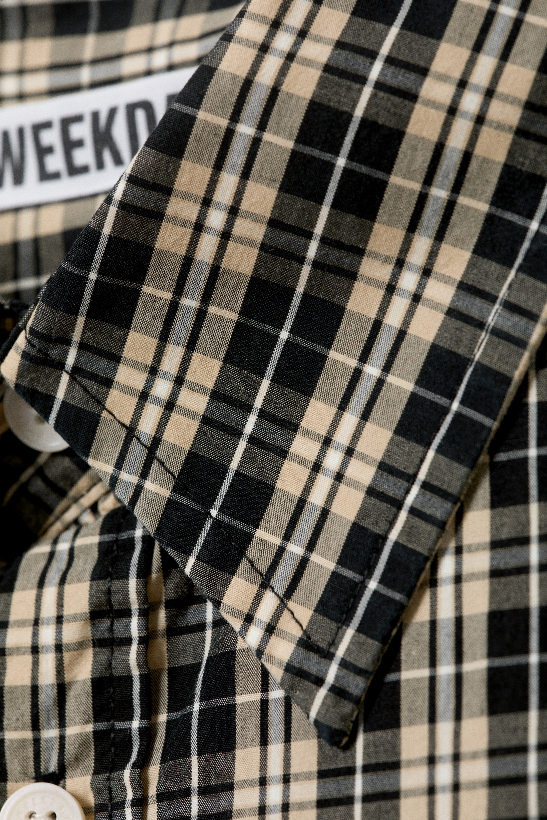 View larger image: A shirt is featuring a black, beige, and white checked pattern. Its pointed collar is visible, along with a white button and a partial brand label displaying 'WEEKD'.