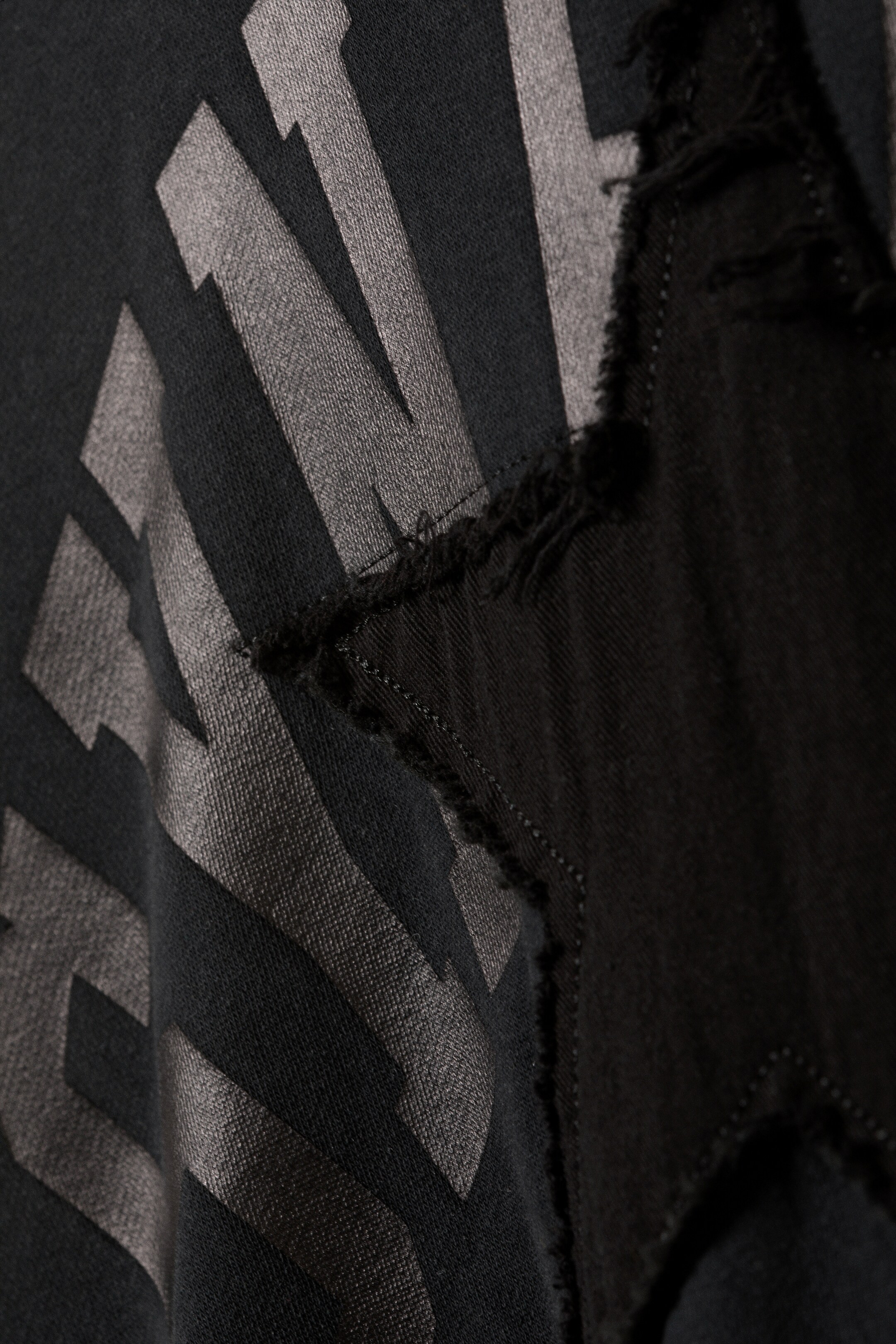 View larger image: The loose-fit graphic hoodie is featuring a bold grey angular print on its dark fabric. A black star-shaped patch is displaying frayed, distressed edges, and it is partially overlapping the graphic.