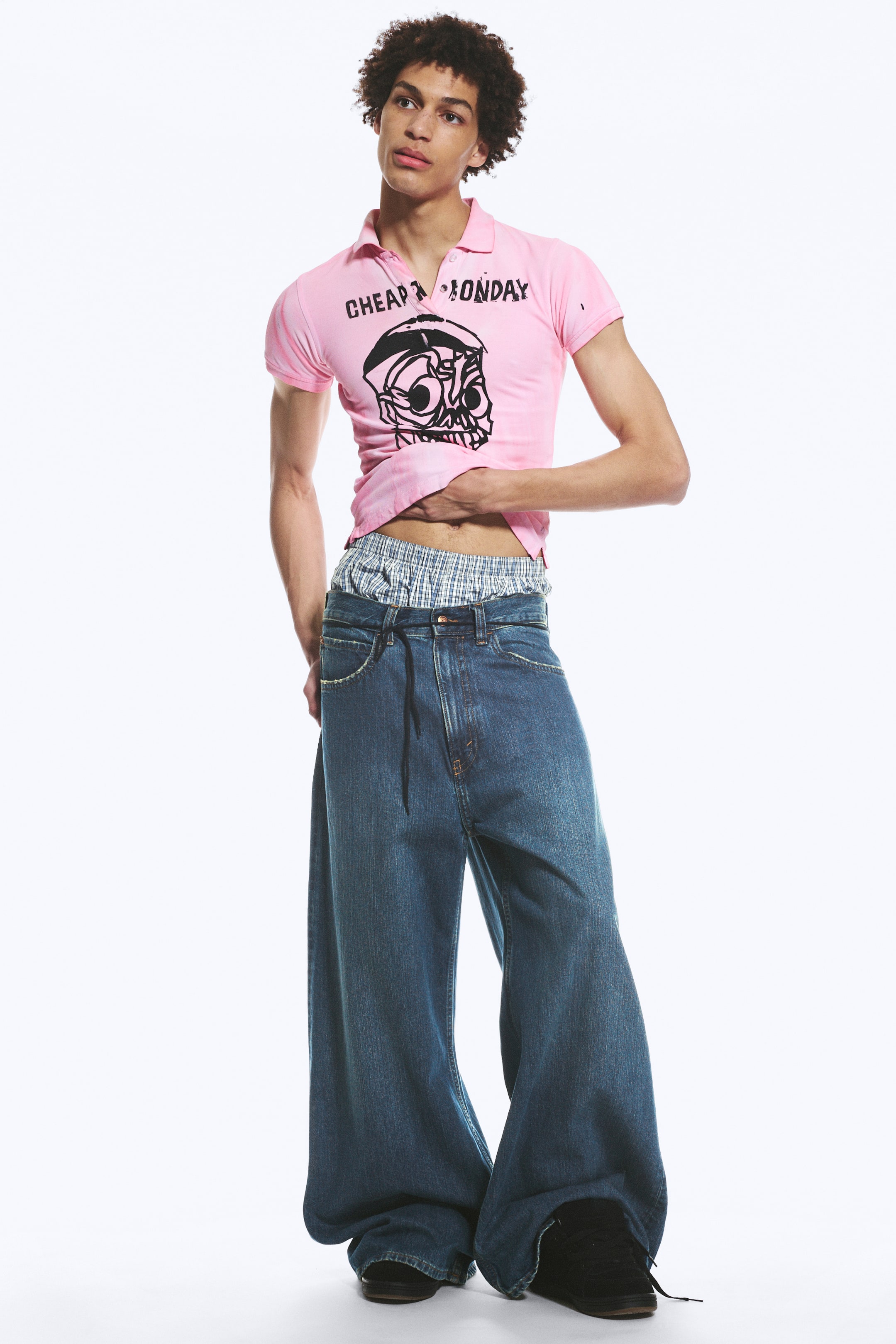 Ver imagen más grande: Se lleva puesta una camiseta polo rosa de manga corta, con el texto "CHEAP MONDAY" en negro y un gráfico de calavera. También se llevan unos vaqueros de mezclilla azul de pernera ancha con cintura ajustable mediante cordón, con bóxers de cuadros visibles por encima de la cintura.