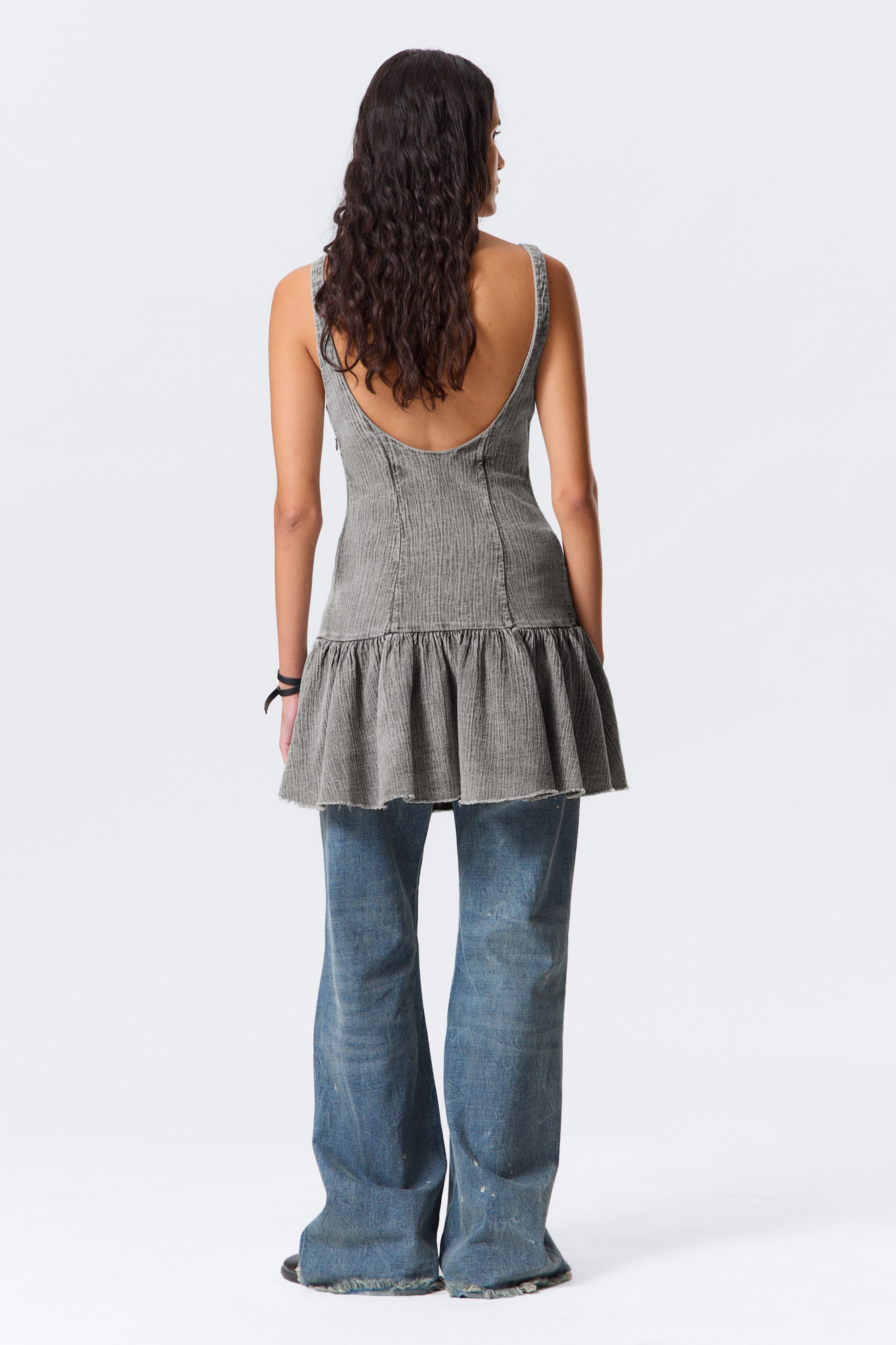 Ingrandisci l'immagine: Un top tunica senza maniche grigio lavato presenta una texture verticale, un corpetto aderente, una profonda scollatura sulla schiena e un orlo raccolto con bordo grezzo. È sovrapposto a jeans destrutturati a gamba larga blu con orli sfilacciati, visti da dietro.