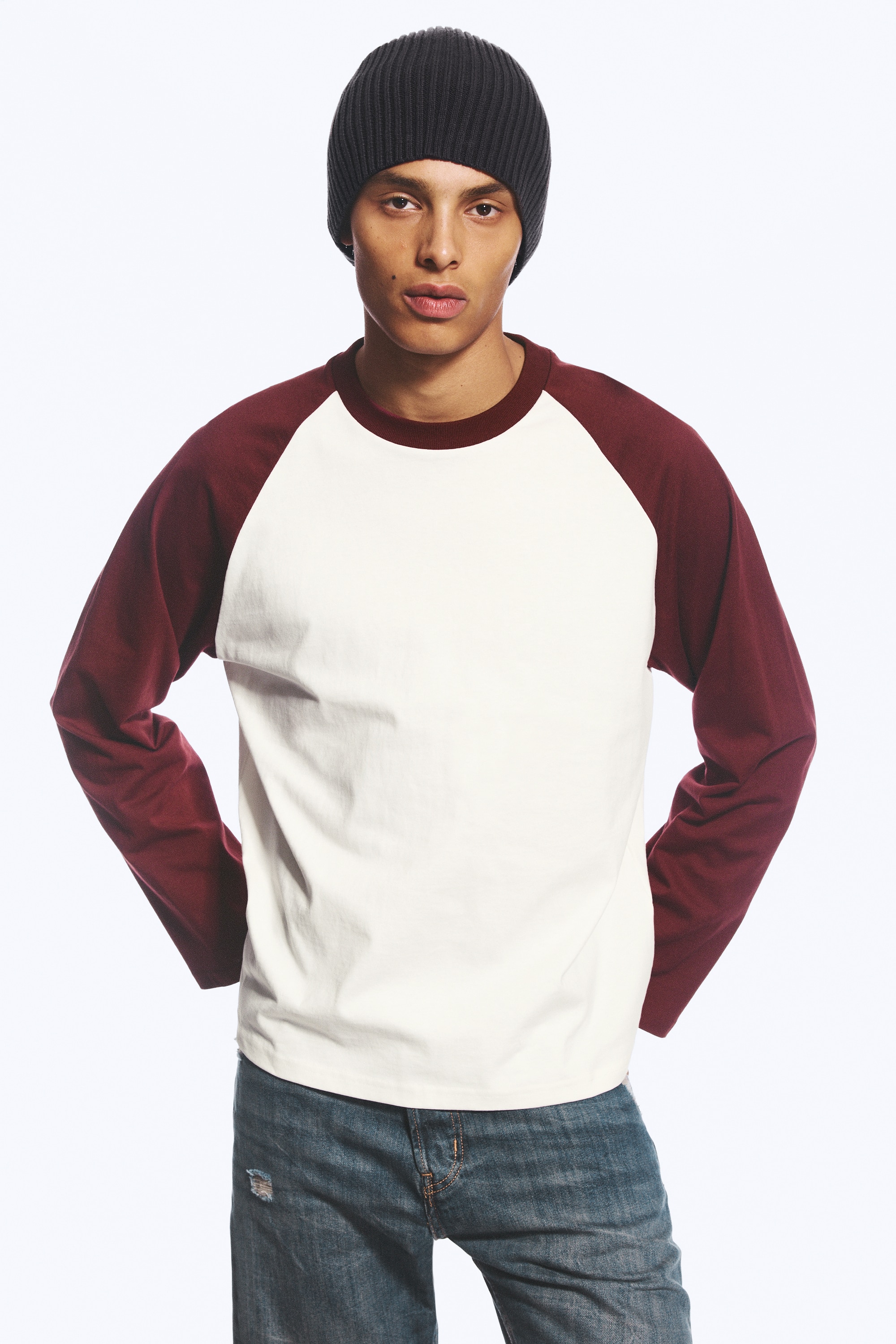 Een raglan-T-shirt met lange mouwen wordt van voren getoond, met een losse, boxy pasvorm en een offwhite body met contrasterende bordeauxrode mouwen. Het T-shirt heeft een bijpassende bordeauxrode geribde ronde hals en recht afgewerkte zomen.
