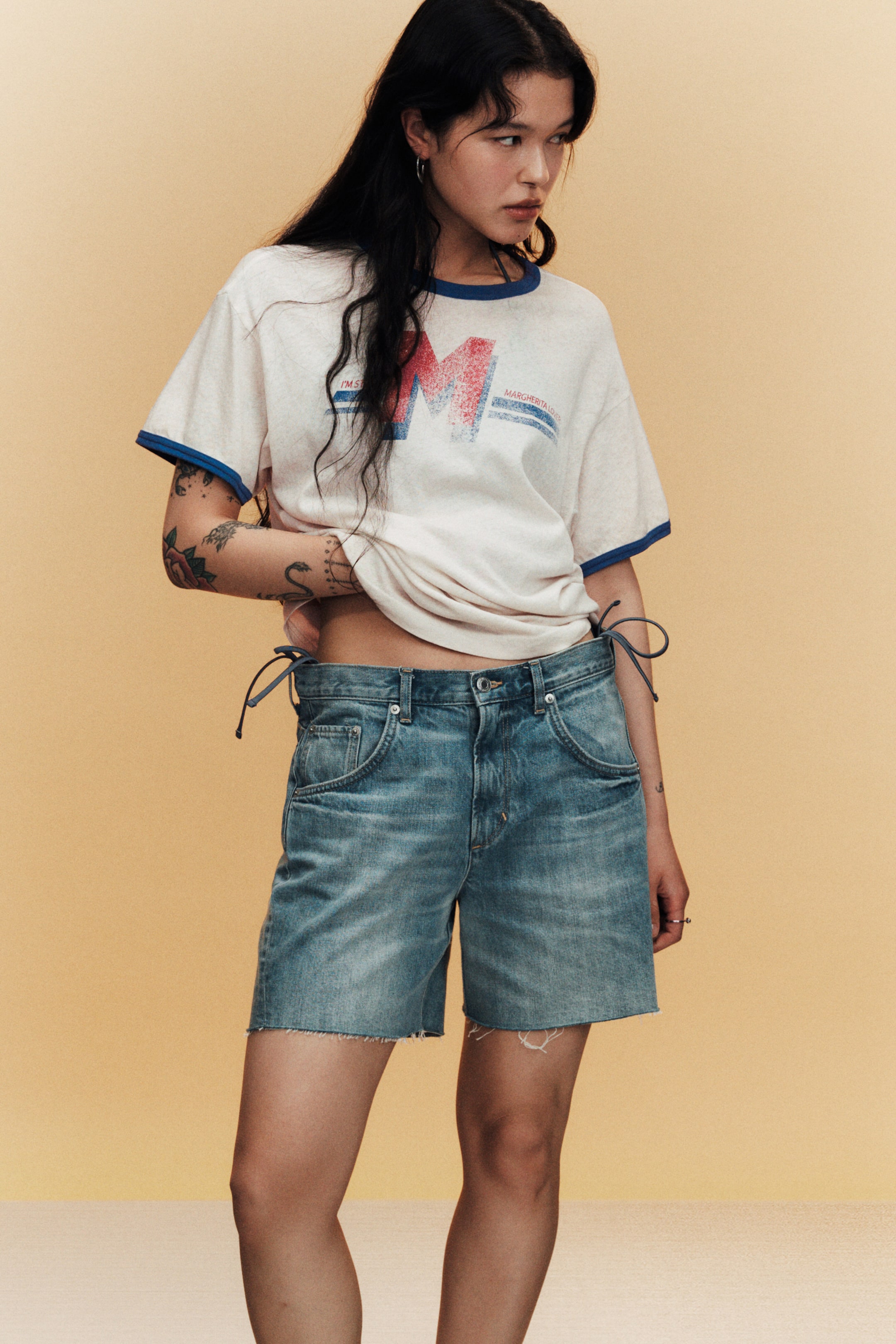 Visualizza immagine più grande: Una t-shirt ringer bianco sporco con bordi blu sul girocollo e sulle maniche corte presenta una grafica 'M' rossa e blu in stile vintage sul petto. I pantaloncini di jeans a lavaggio chiaro offrono una vestibilità rilassata, un orlo grezzo e lacci blu che stringono i lati della cintura.