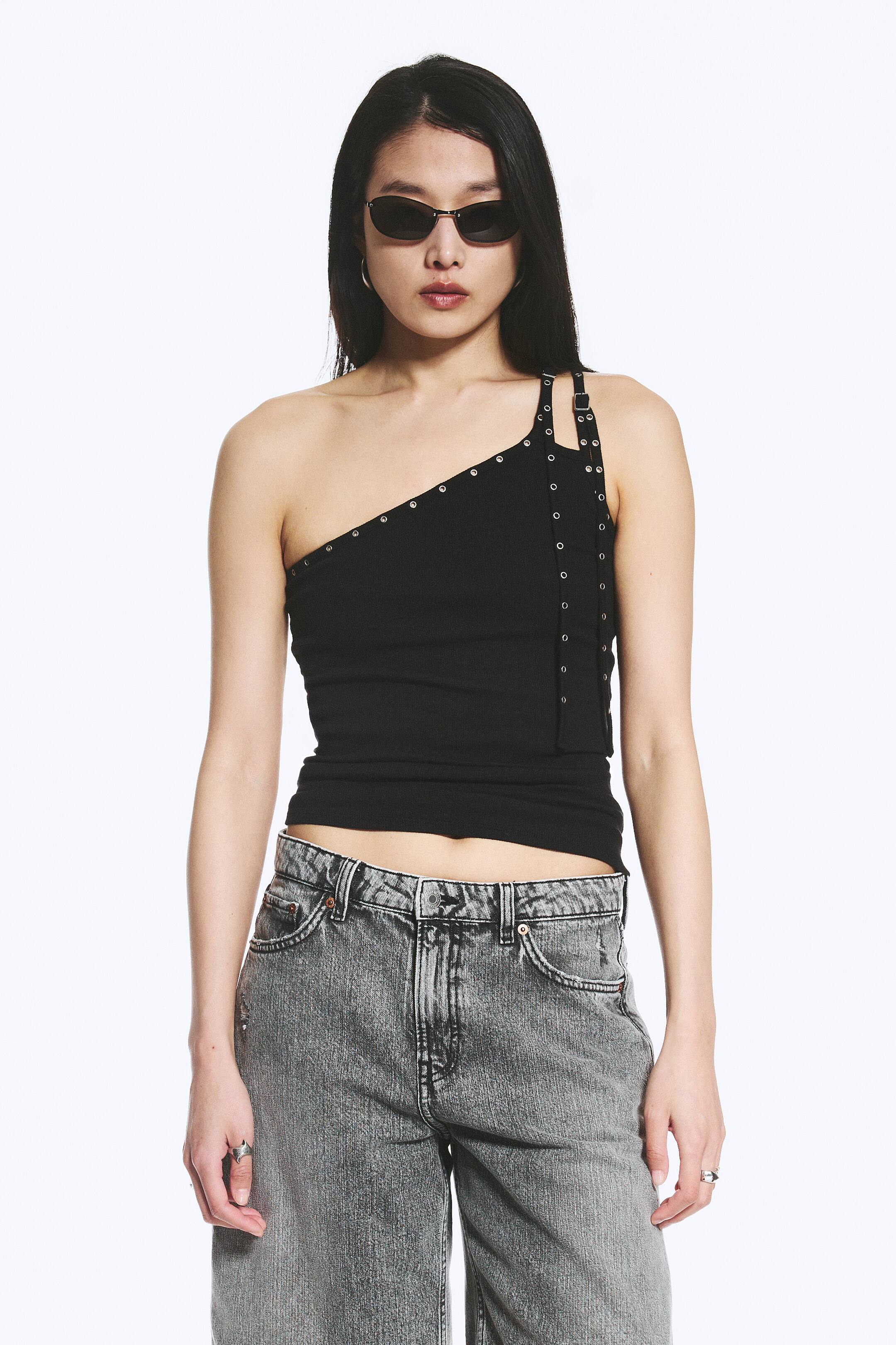 Agrandir l'image: Un débardeur noir ajusté présente un col asymétrique, une coupe sans manches, des œillets en métal argenté et des bretelles doubles allongées avec des détails de boucle sur une épaule. Le modèle porte le débardeur.