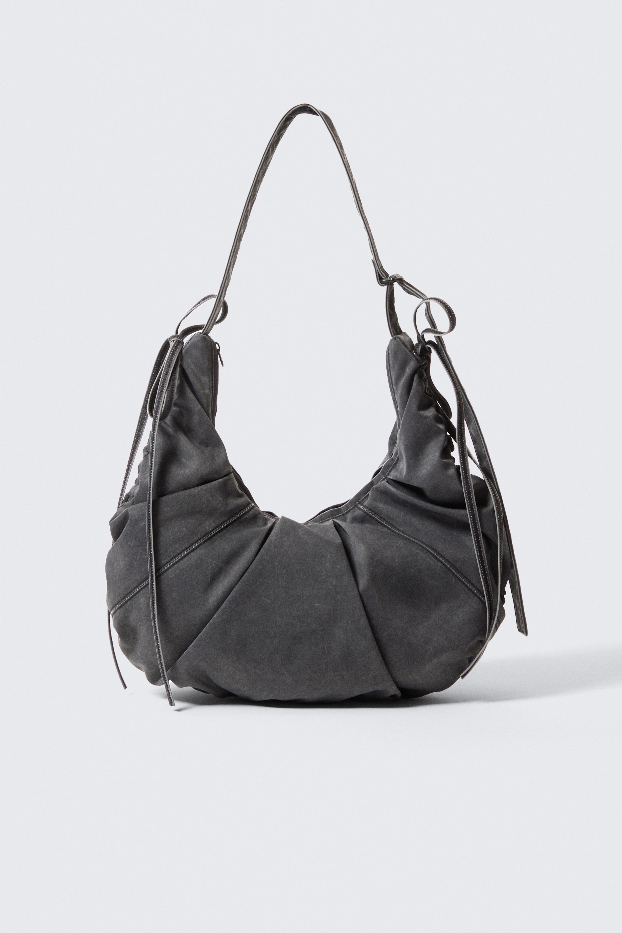 Grösseres Bild ansehen: Eine gewaschene, kohlefarbene, halbmondförmige Handtasche wird ausgestellt, die eine distressed Textur und strahlende Falten über ihren weichen, voluminösen Körper aufweist. Ein dünner Schulterriemen erhebt sich aus den oberen Öffnungen, die mit sichtbaren Bändern zusammengezogen sind.