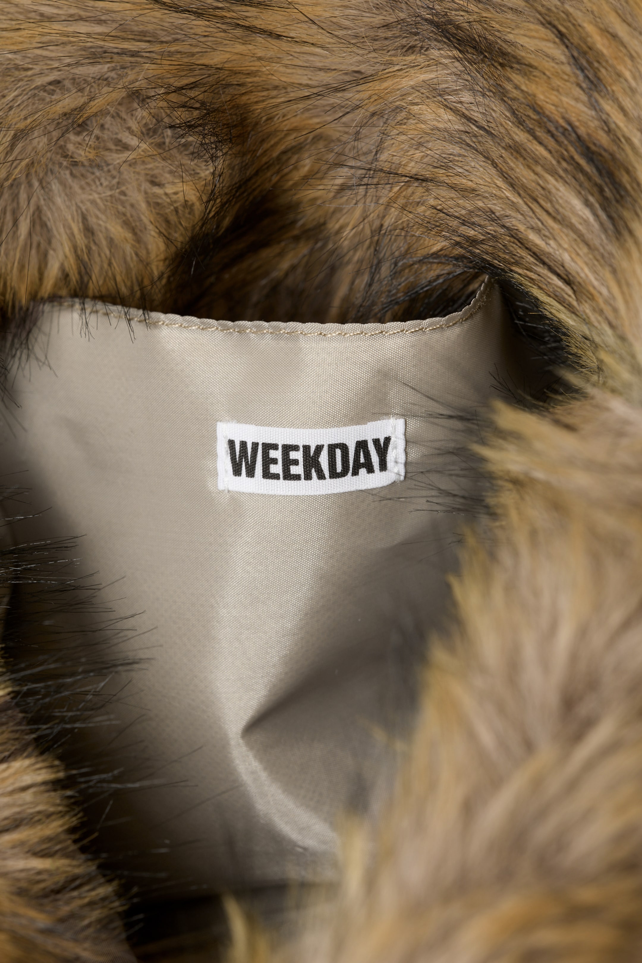 Größeres Bild ansehen: Das hellgraue Innenfutter eines Kleidungsstücks ist von braunem und schwarzem Kunstfell umrandet. Ein weißes gewebtes Etikett mit der Aufschrift "WEEKDAY" in schwarzem Text ist auf das Futter genäht.
