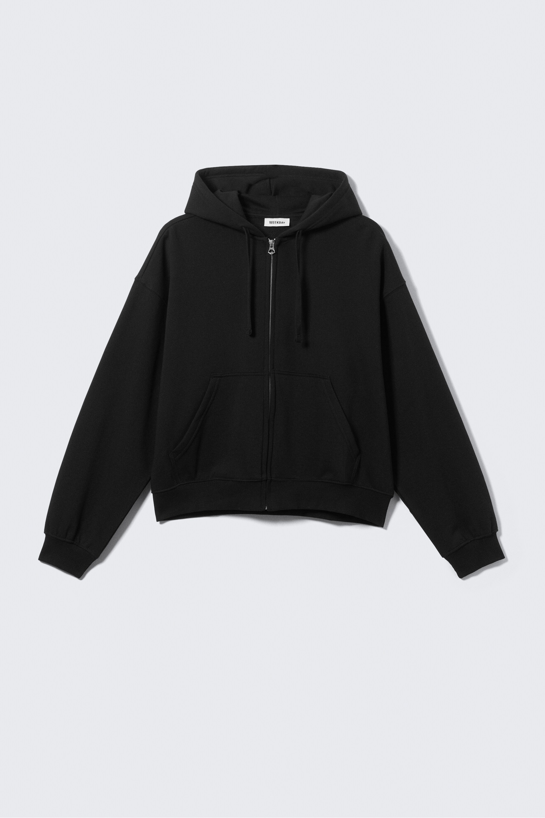View larger image: A black zip-up hoodie is presenting front-facing, featuring a silver-toned front zipper and a large hood. It is including a kangaroo pocket, dropped shoulders, and ribbed cuffs and hem. A small white 'WEEKDAY' label is visible on the inside of the neck.