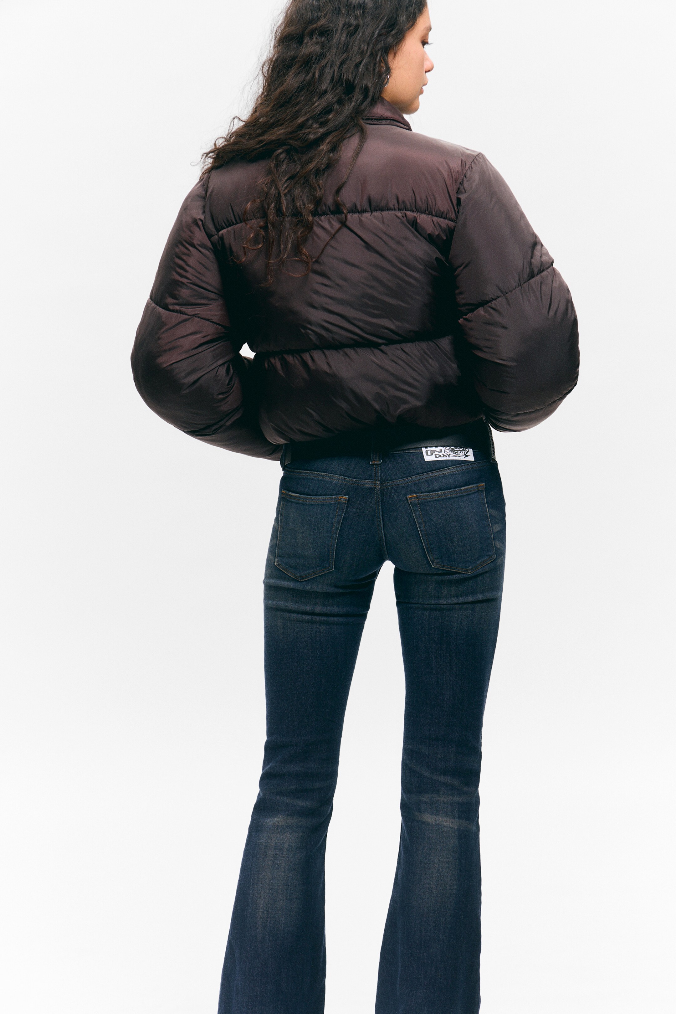 View larger image: A dark brown cropped puffer jacket is being shown from the back, featuring puffed sections. It is styled with dark wash flared jeans, displaying back pockets and a small white label on the right side of the waistband.