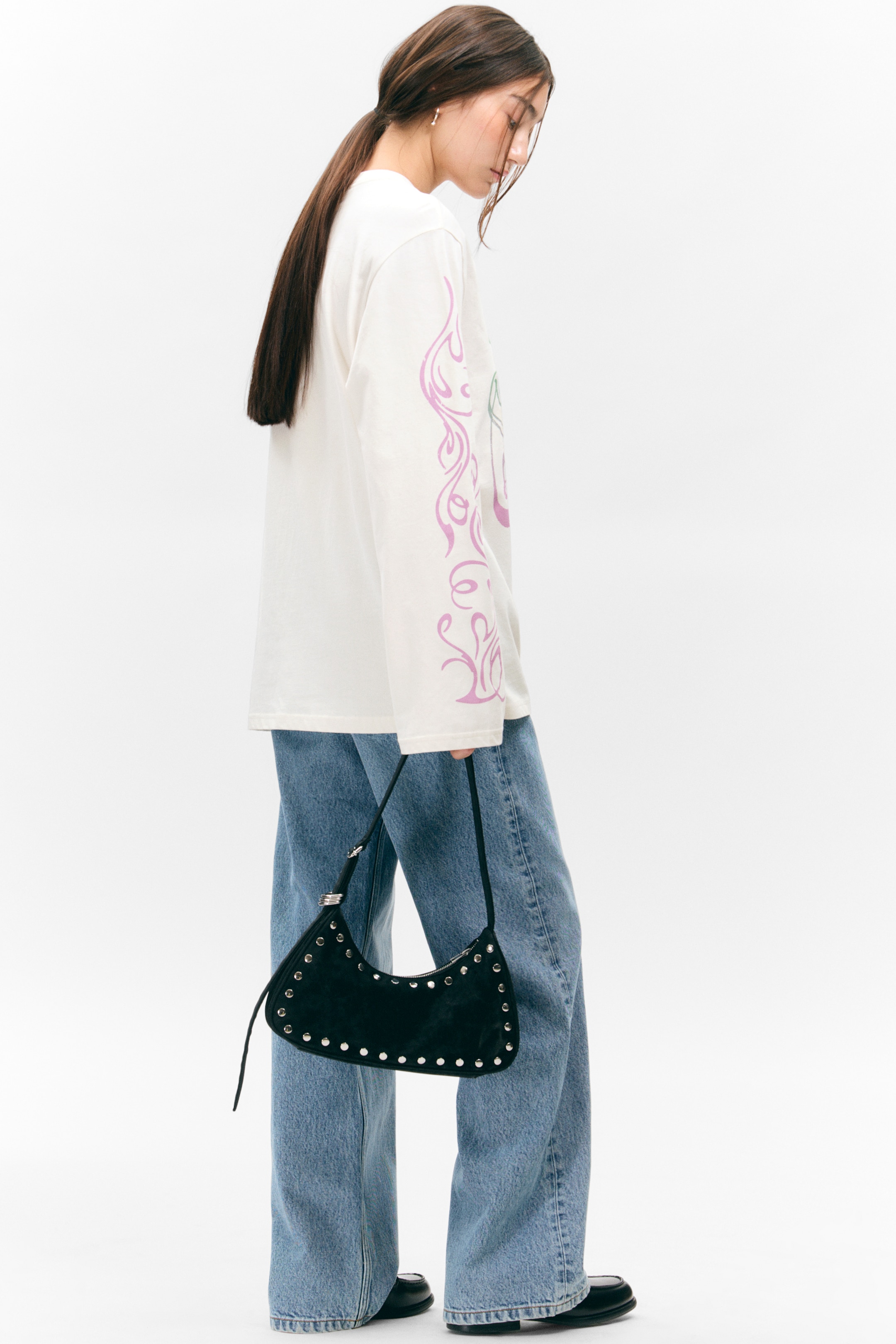 View larger image: An off-white long-sleeved T-shirt, featuring a relaxed fit, crew neckline, dropped shoulders, and wide sleeves with lavender graphic prints, is being worn by the model. The model is styling the T-shirt with light blue wide-leg jeans and is carrying a black studded shoulder bag.