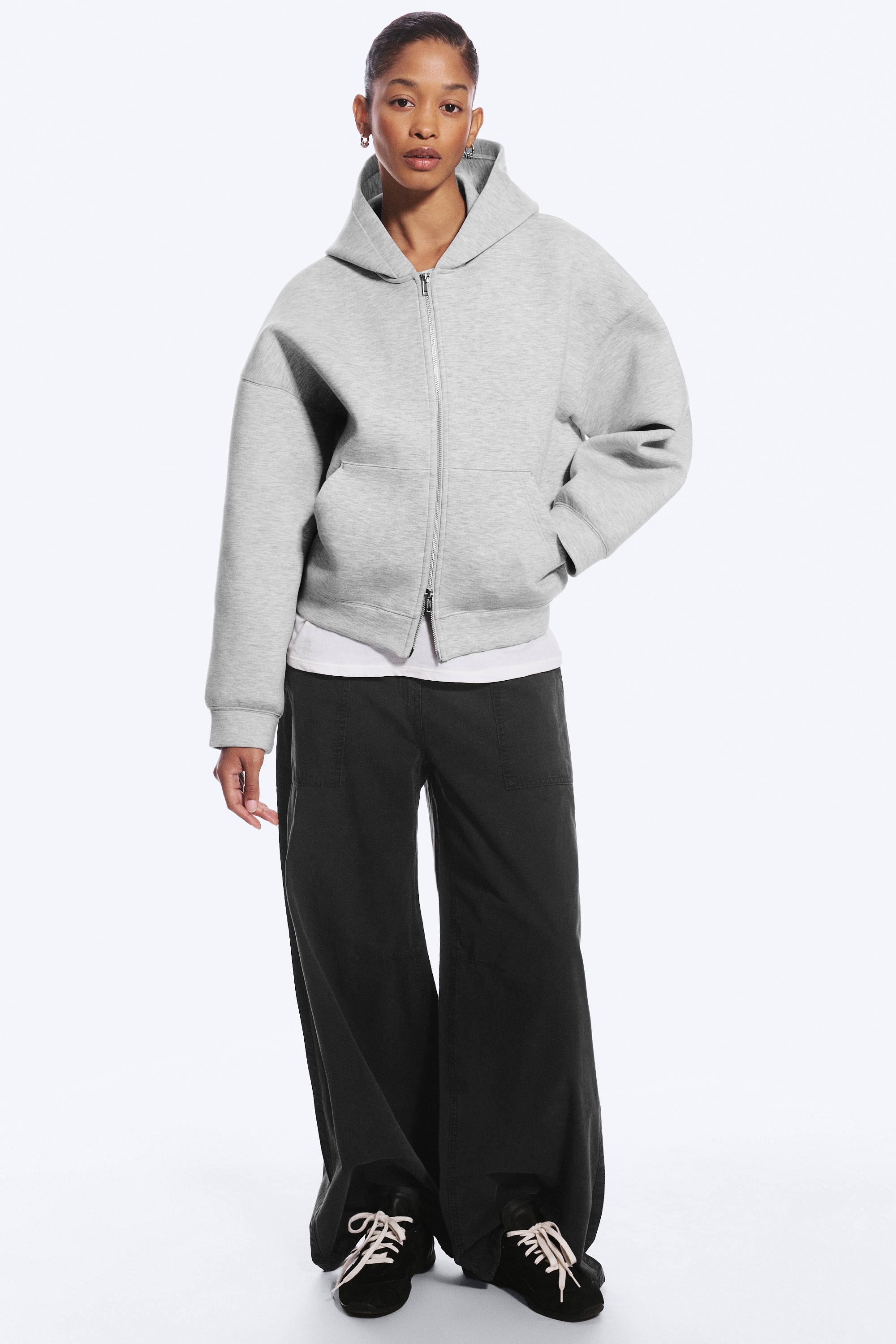 Een lichtgrijze, oversized hoodie met rits ligt plat en wordt van voren getoond, waarbij de gestructureerde pasvorm met verlaagde schouders zichtbaar is. Het kledingstuk heeft een dubbellaagse capuchon, een zilverkleurige tweewegrits aan de voorkant, gespleten steekzakken en omgeslagen boorden aan de mouwen en zoom.