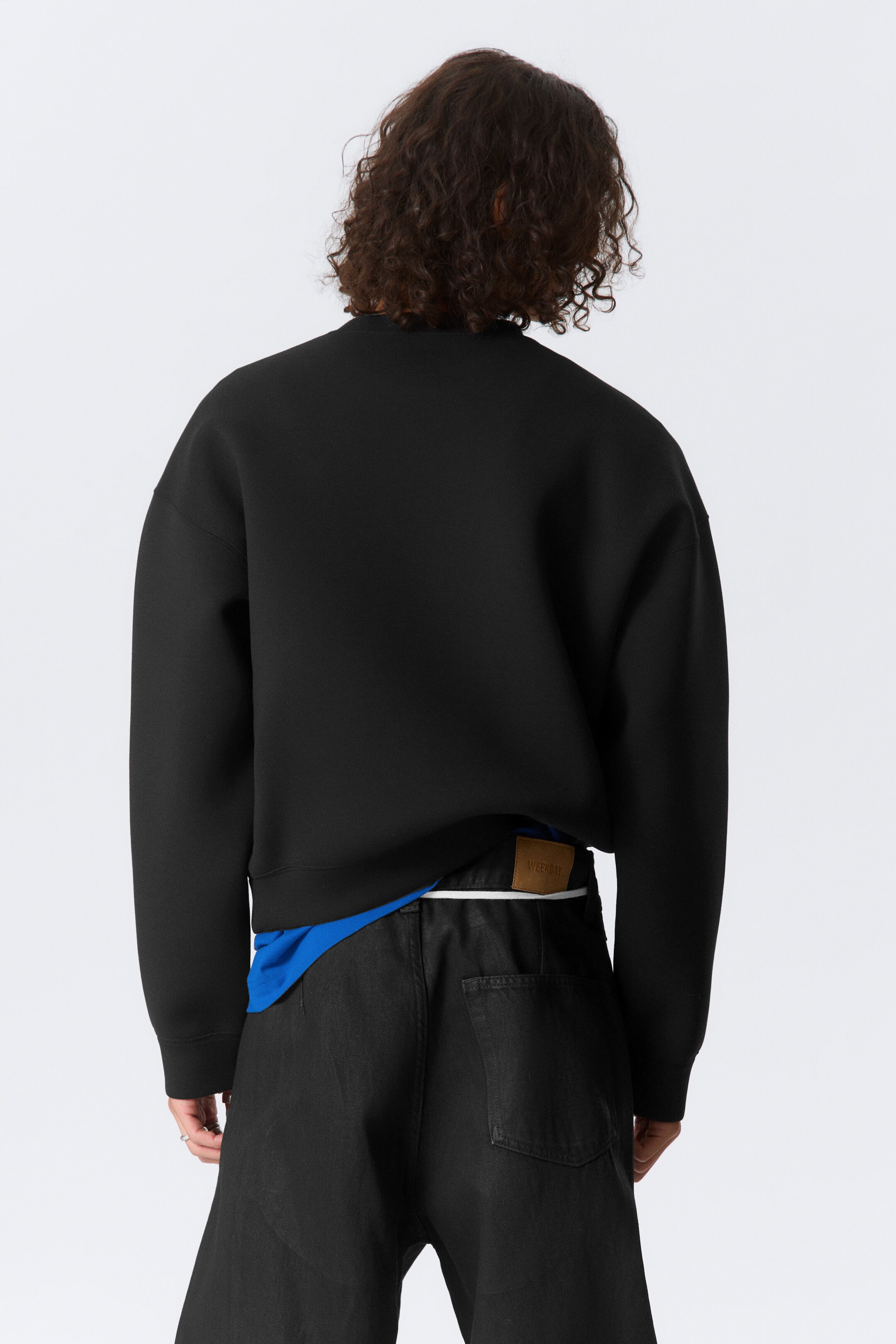Se større billede: En sort sweatshirt bliver præsenteret bagfra, med et glat, struktureret stof, lange ærmer og en afslappet pasform. Dens kant hviler over taljelinjen af sorte bukser, med en blå t-shirt synlig nedenunder.