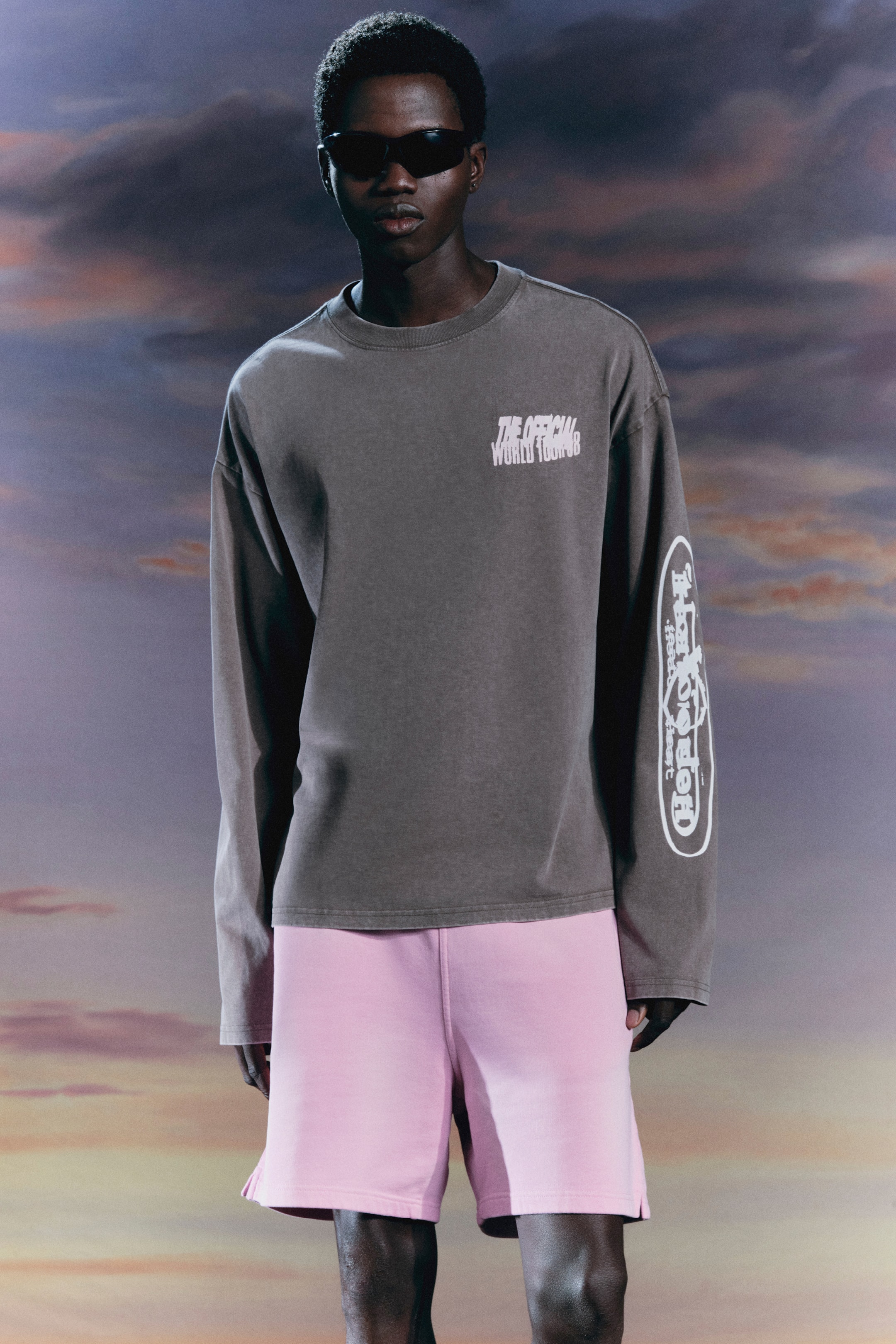 Visa större bild: En mörkgrå långärmad t-shirt med ett vitt "THE OFFICIAL WORLD TOUR '98"-tryck på bröstet och ett förlängt vitt tryck på vänster ärm. Rosa shorts med en avslappnad passform och små sidoslitsar vid fållen.