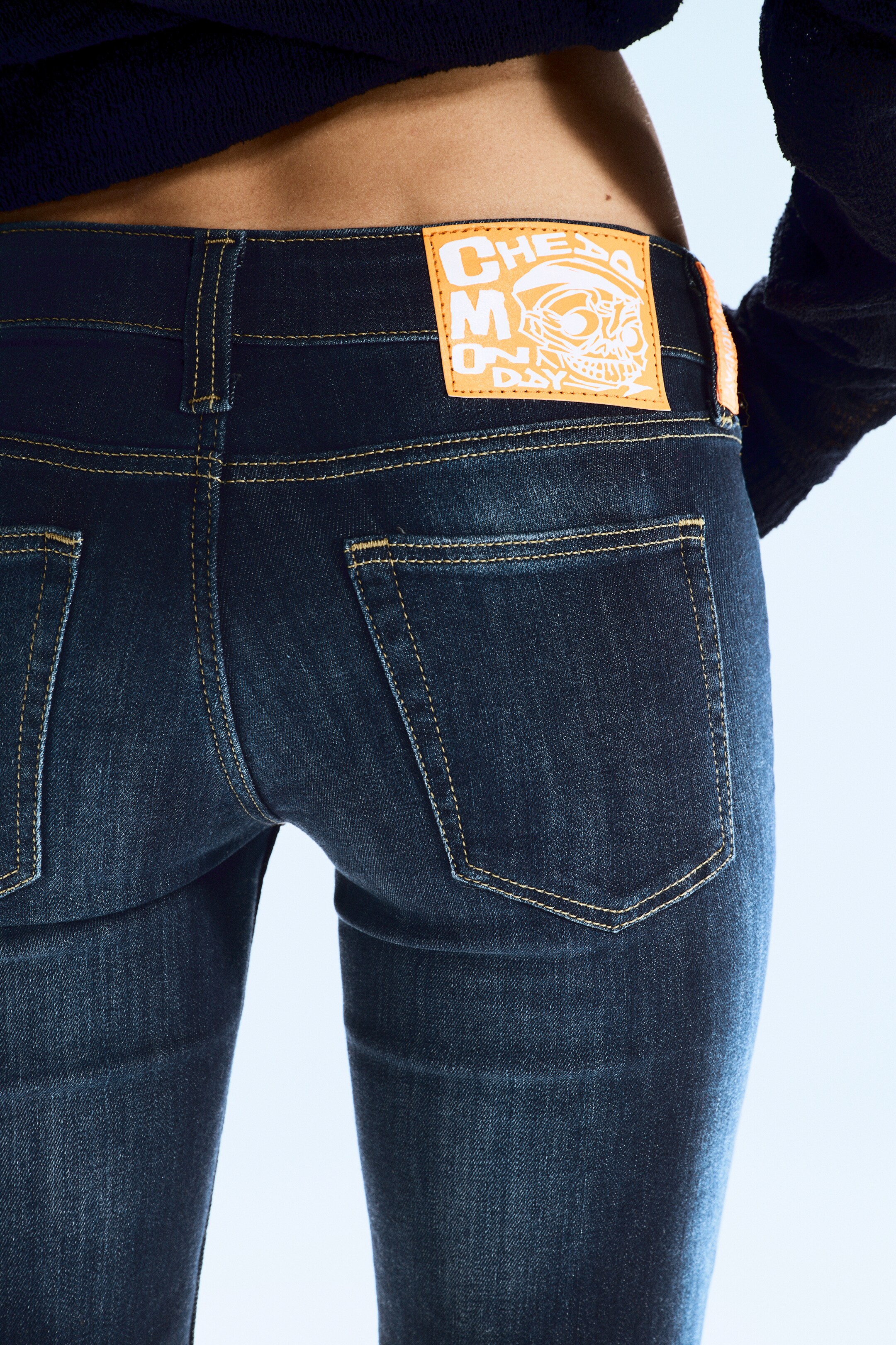Ingrandisci l'immagine: Jeans slim-fit a lavaggio scuro sono presentati dal retro, leggermente angolati a sinistra. Sono caratterizzati da due tasche posteriori applicate con cuciture gialle a contrasto, e una vivace etichetta del marchio arancione, con un disegno di teschio bianco e il testo "CHEAP MONDAY", è esposta sulla cintura.