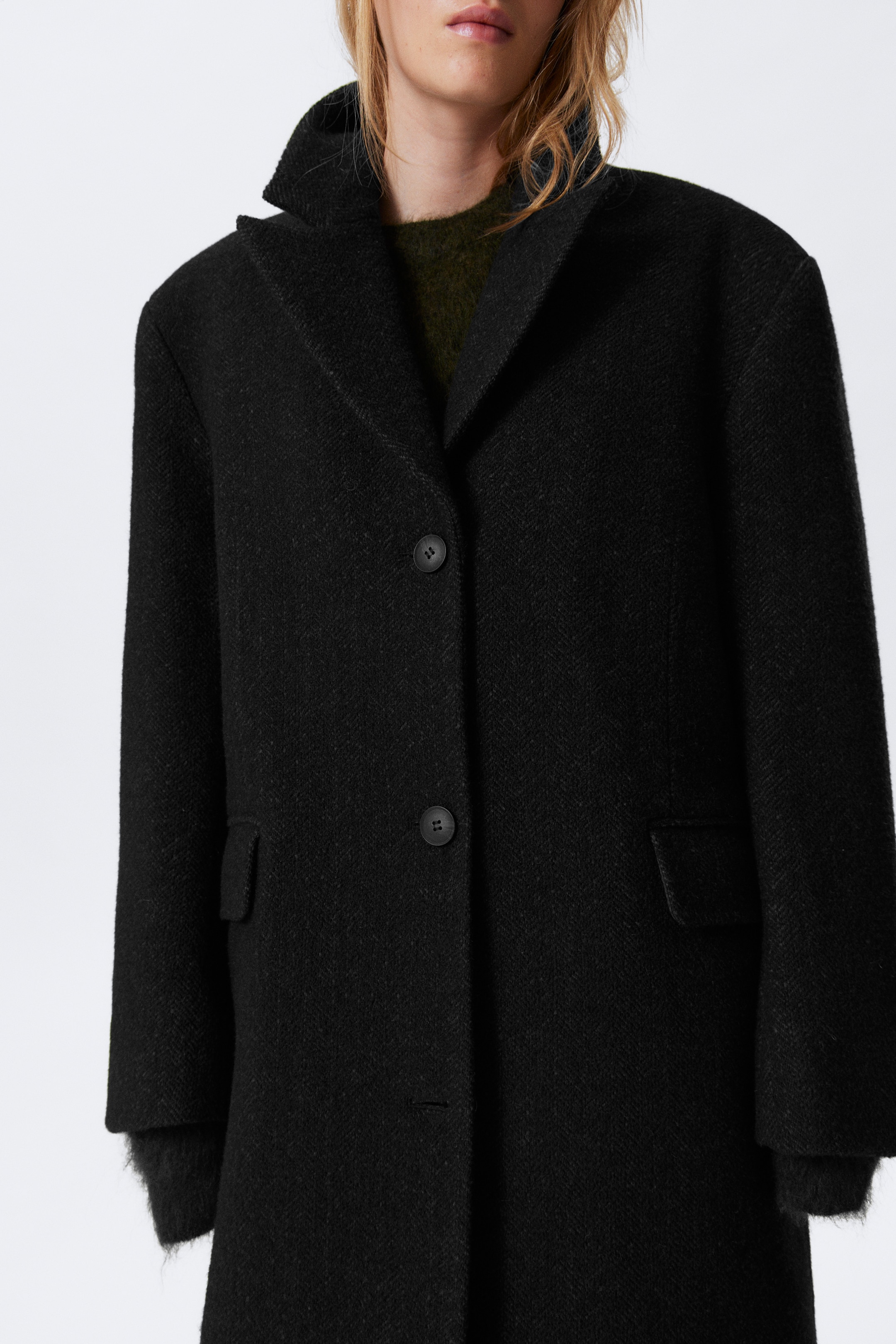 View larger image: A black textured coat is presented from a slightly angled front view, featuring notch lapels, a two-button closure, and two flap pockets. A dark green sweater is being worn underneath, with its cuffs extending from the coat's sleeves.